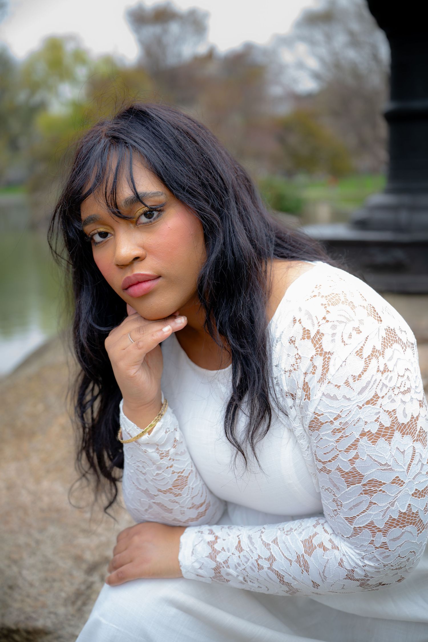 Young woman in white lace dress poses thoughtfully near pond in park.
