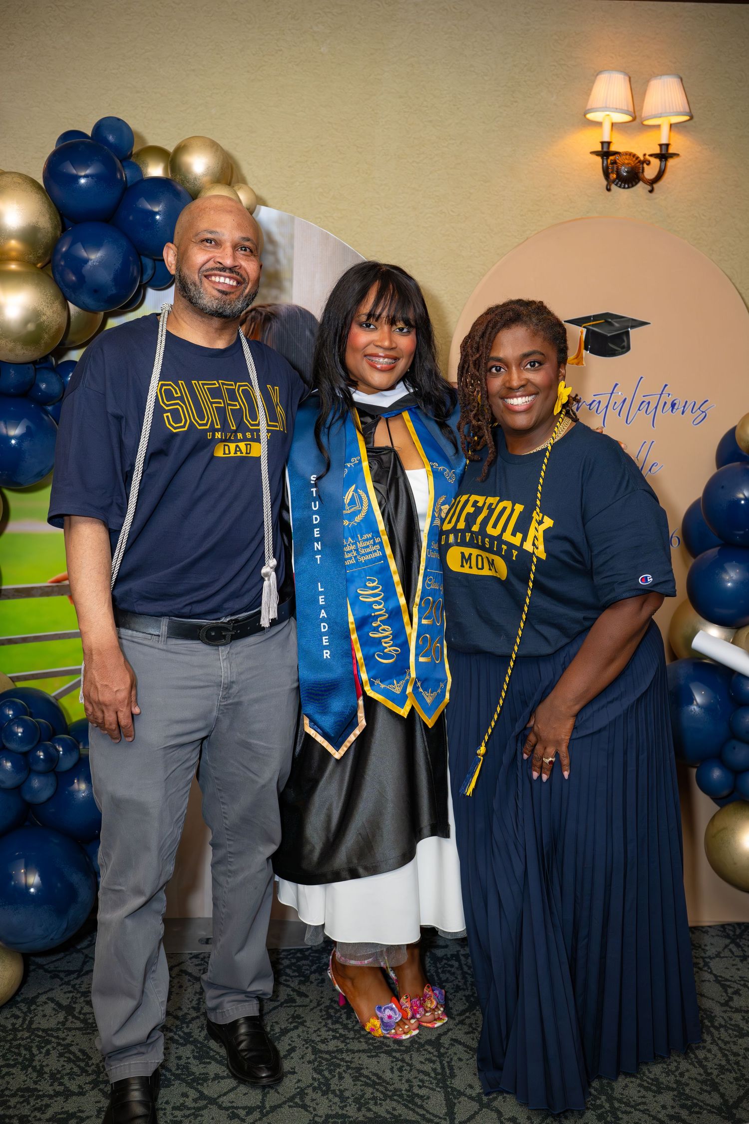 Suffolk University graduates pose with proud parents wearing Suffolk Dad and Mom shirts at a graduation celebration.