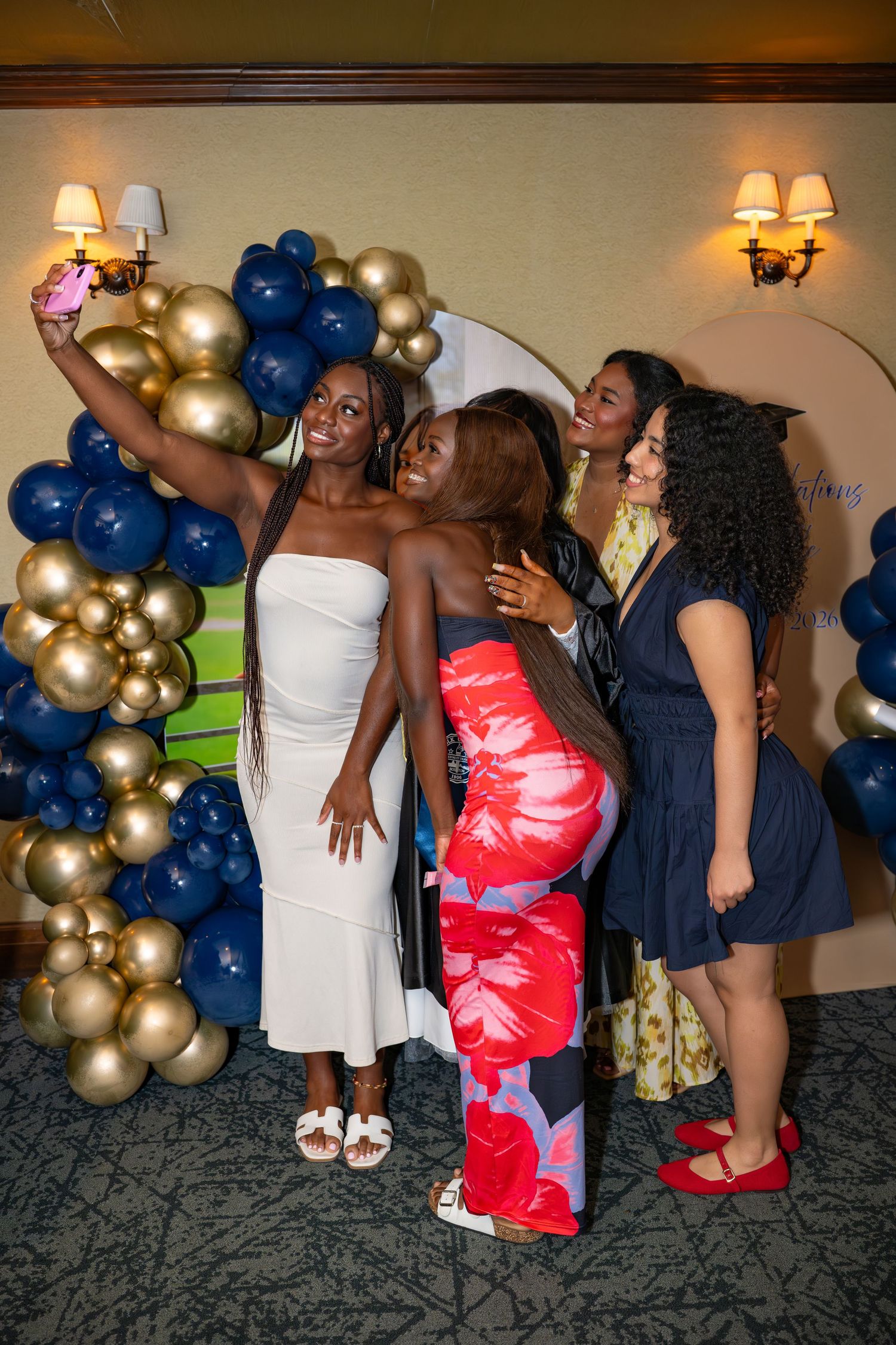 Four women pose for a selfie in front of navy blue and gold balloon decorations at a formal event.