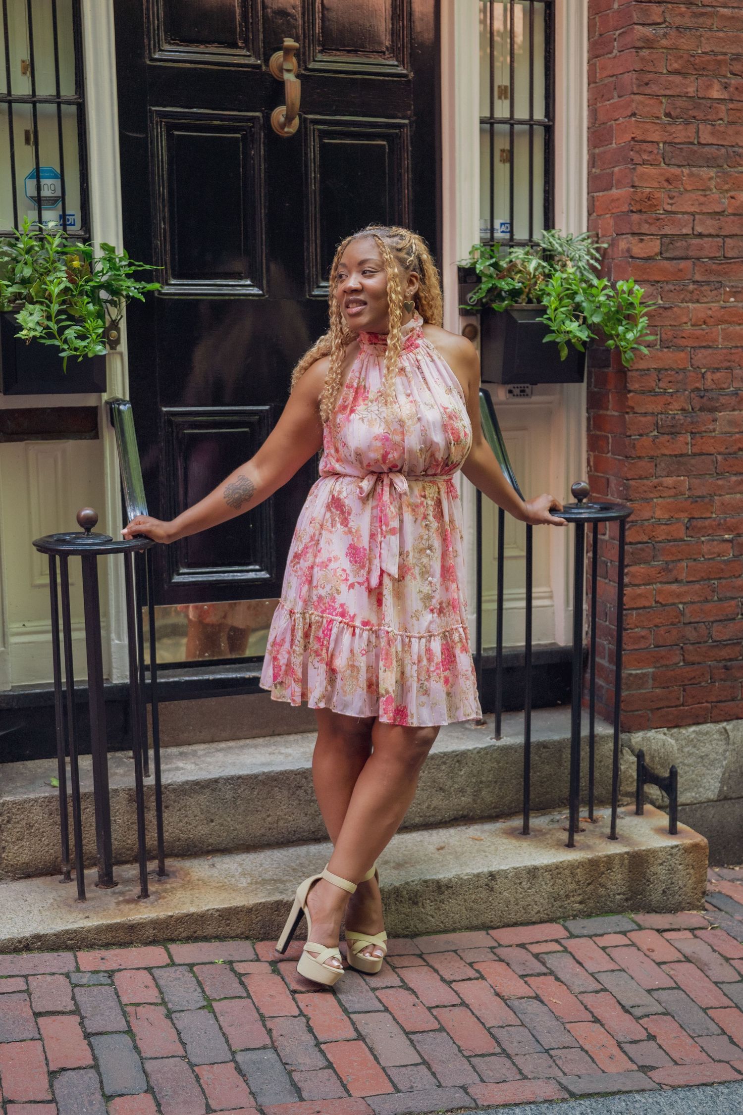 Woman in pink floral halter dress smiles on brick steps outside elegant dark door with potted plants.