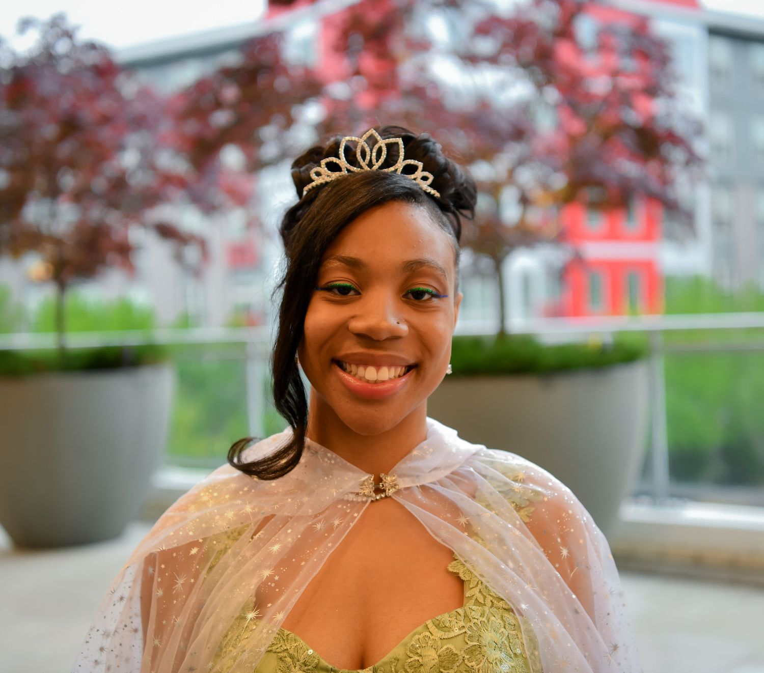 Smiling young woman in gold princess dress and tiara wearing sheer cape outdoors.