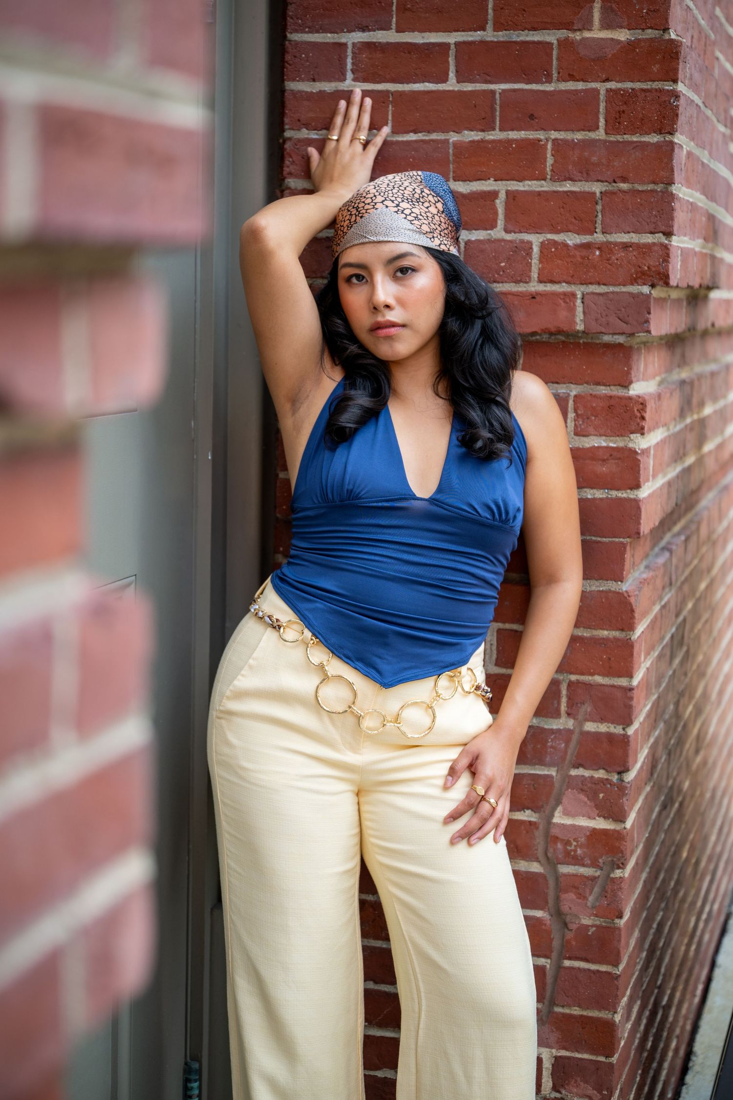 Woman in blue halter top, cream wide-leg pants, gold chain belt, and headscarf poses against brick wall.