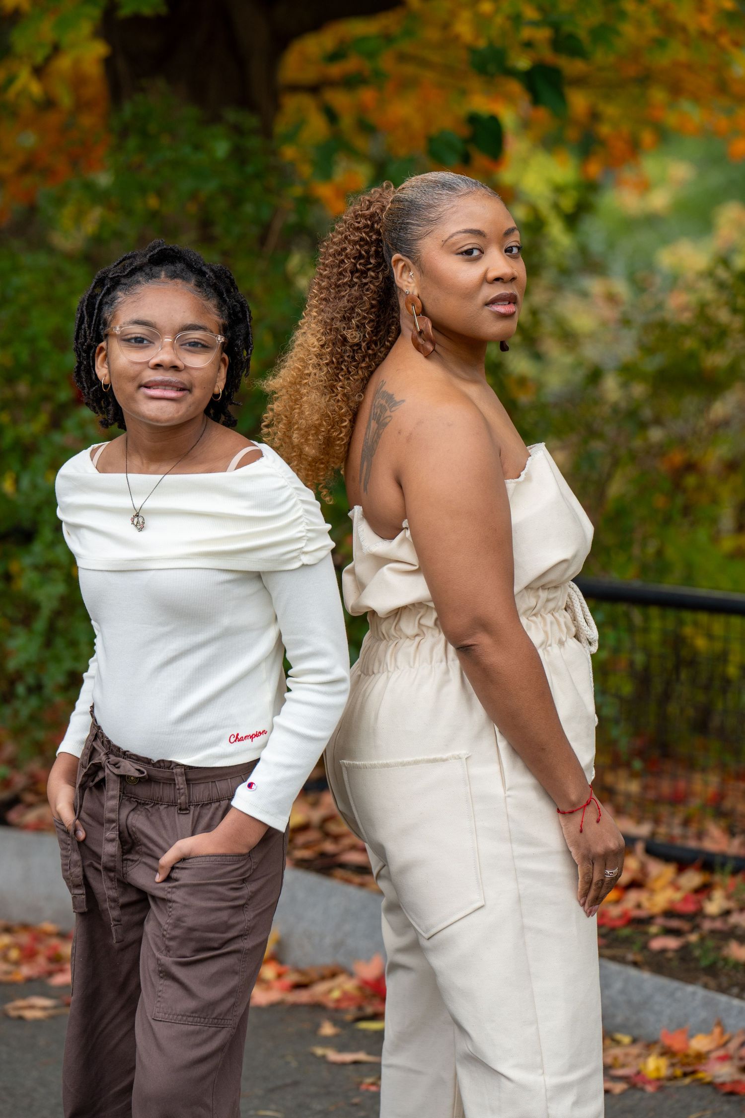 Two women pose outdoors among colorful autumn foliage, one in a white off-shoulder top and brown pants, the other in a cream jumpsuit.