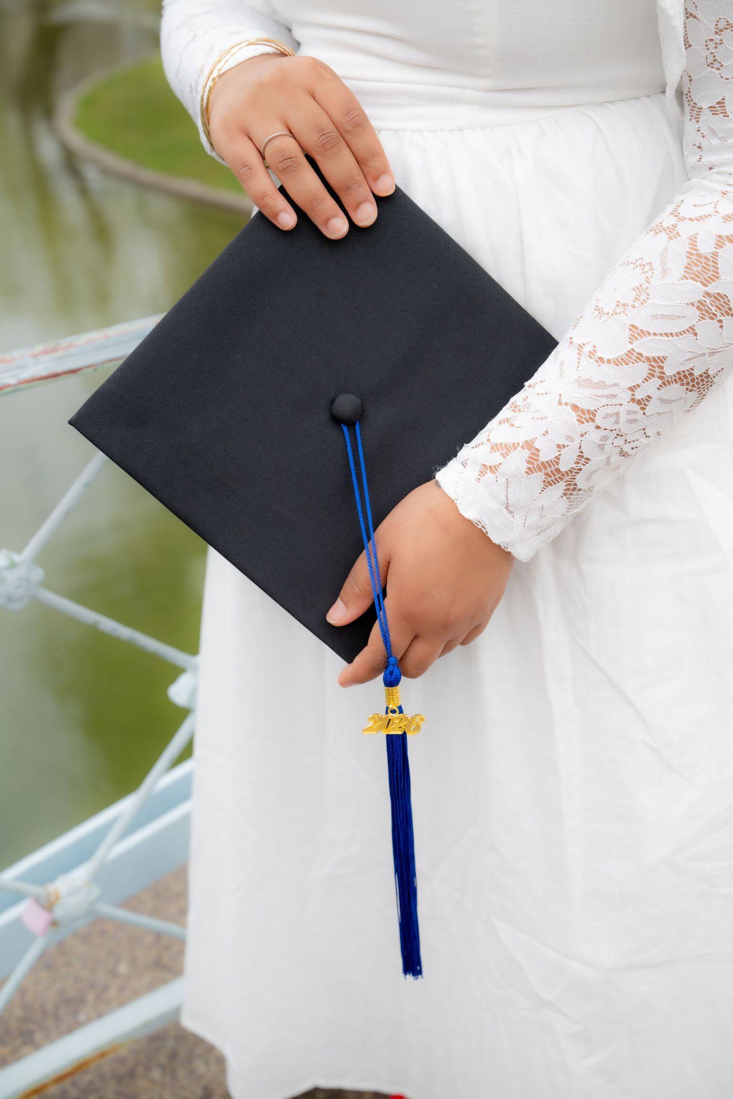 Graduate in white lace dress holding black mortarboard cap with blue tassel outdoors.