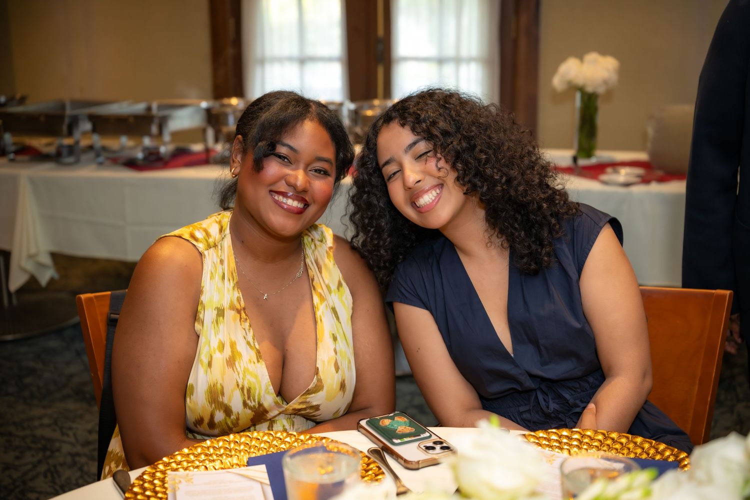 Two smiling women pose at decorated table during indoor graduation celebration party event.