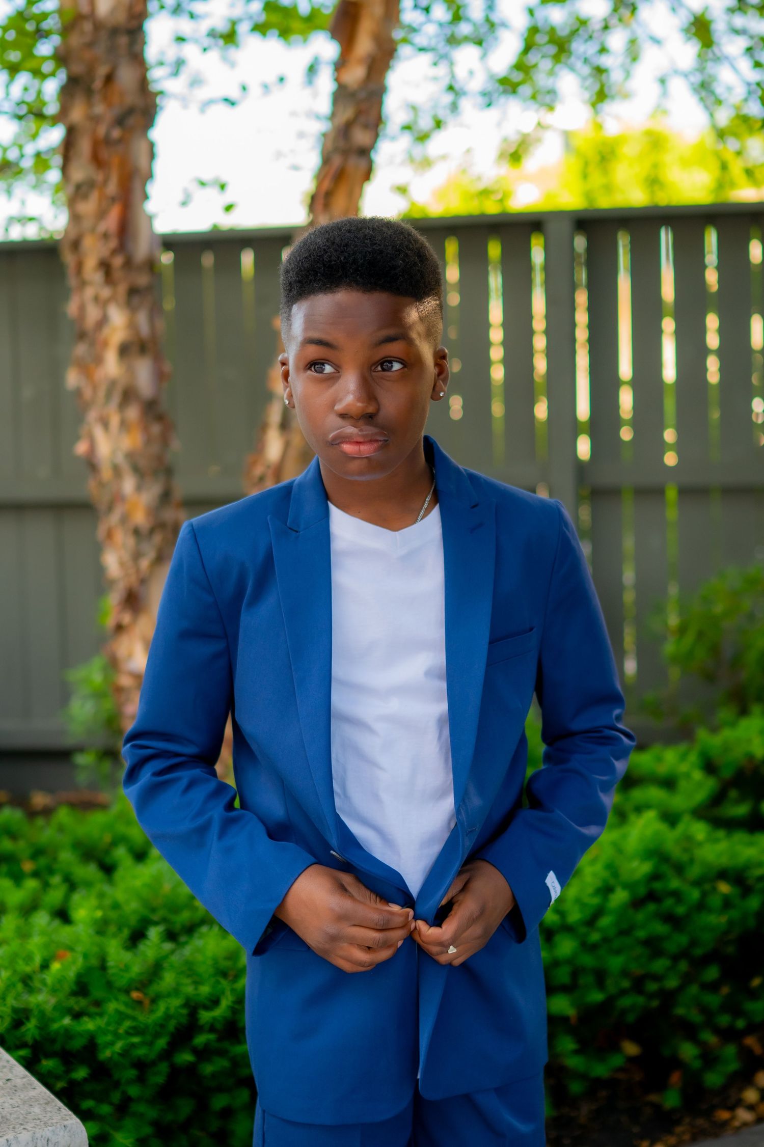 Young man in blue blazer and white shirt posing outdoors in garden setting.