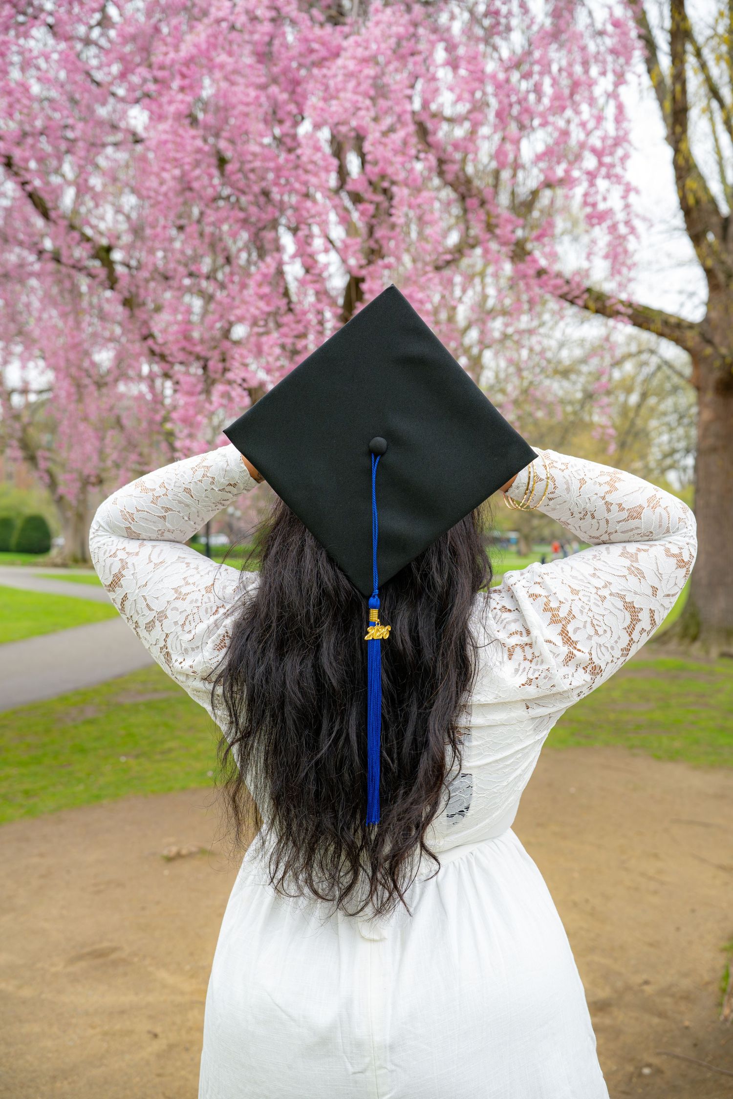 A graduate in a white lace dress adjusts her black mortarboard cap while standing before blooming pink cherry blossom trees.