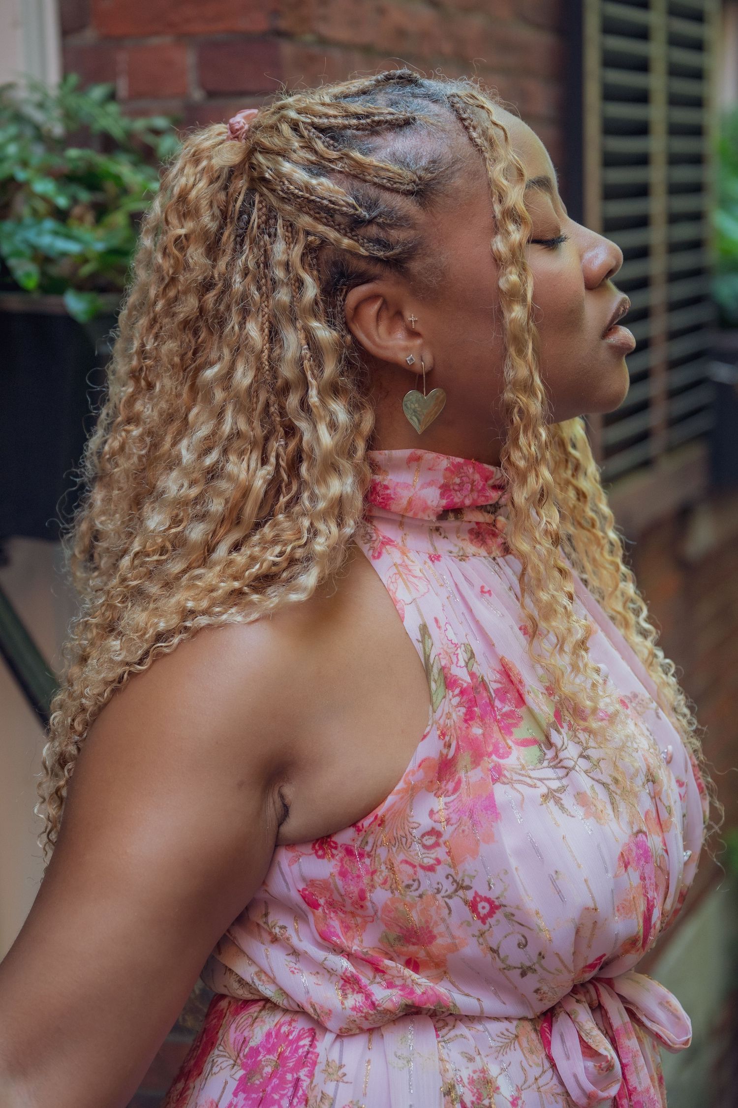 A woman with long blonde braided curly hair wearing a pink floral halter dress poses outdoors near a brick wall.