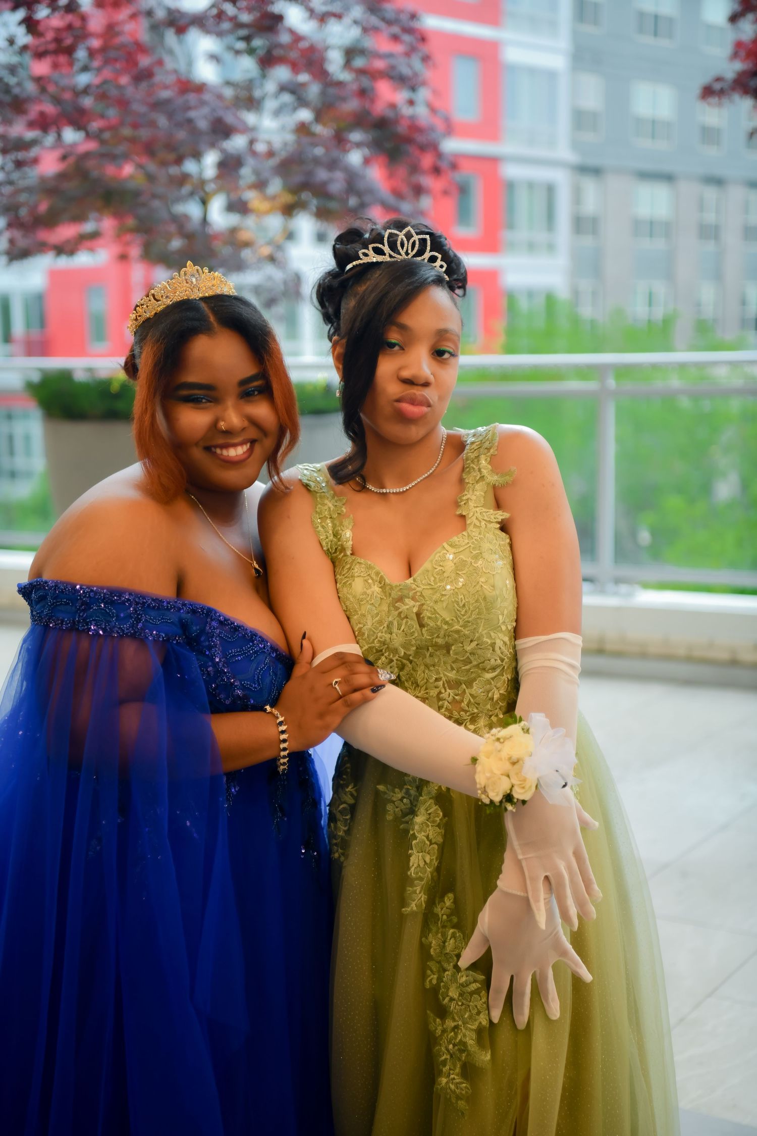 Two young women in formal prom gowns and tiaras pose together outdoors in blue and gold dresses.
