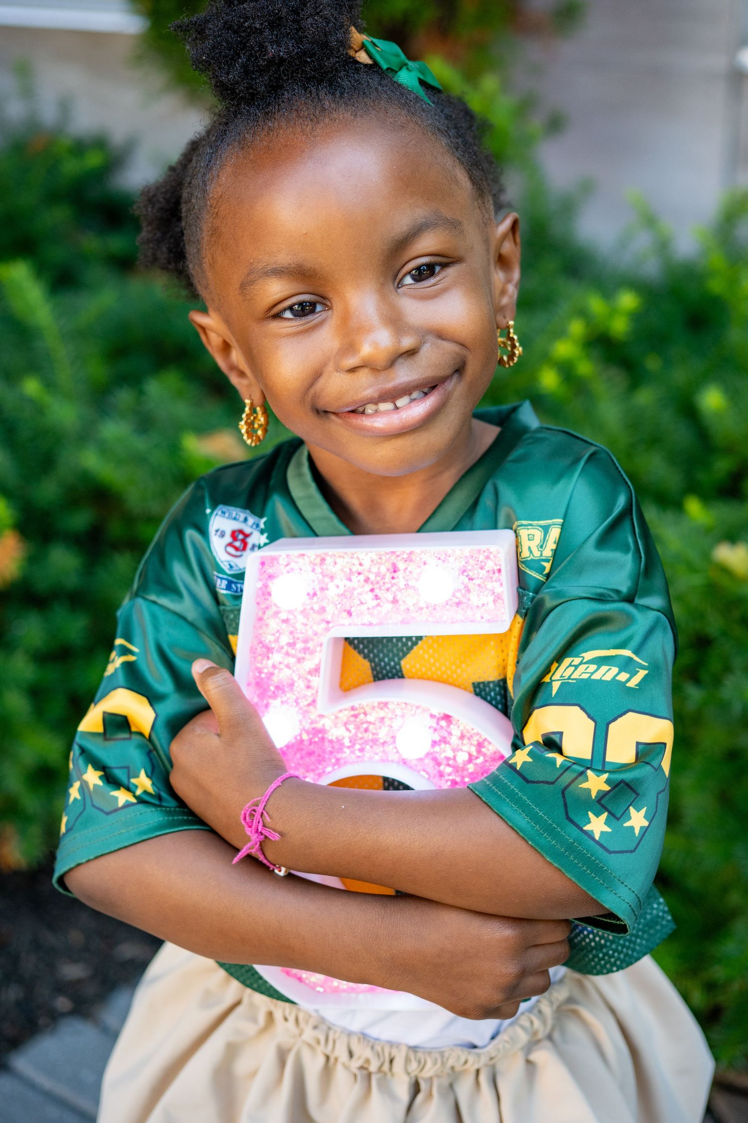 Smiling girl in green jersey hugs glittery pink number five outdoors.
