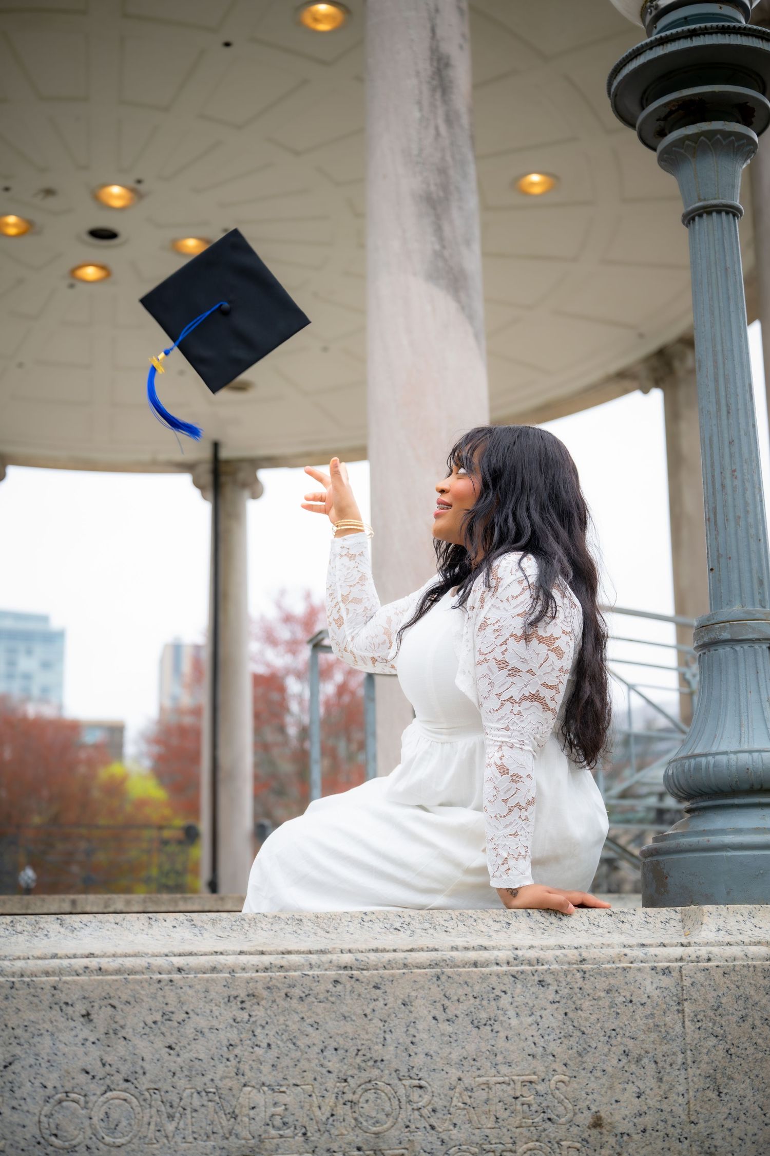 Young woman in white lace dress tossing graduation cap outdoors near decorative columns, celebrating academic achievement.