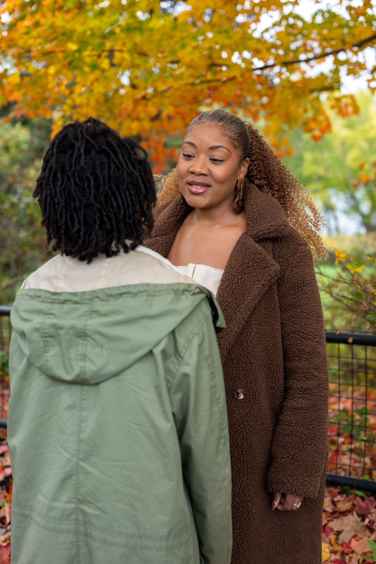 Two women talk outdoors among vibrant orange autumn leaves, one in a brown teddy coat and one in a green hooded jacket.