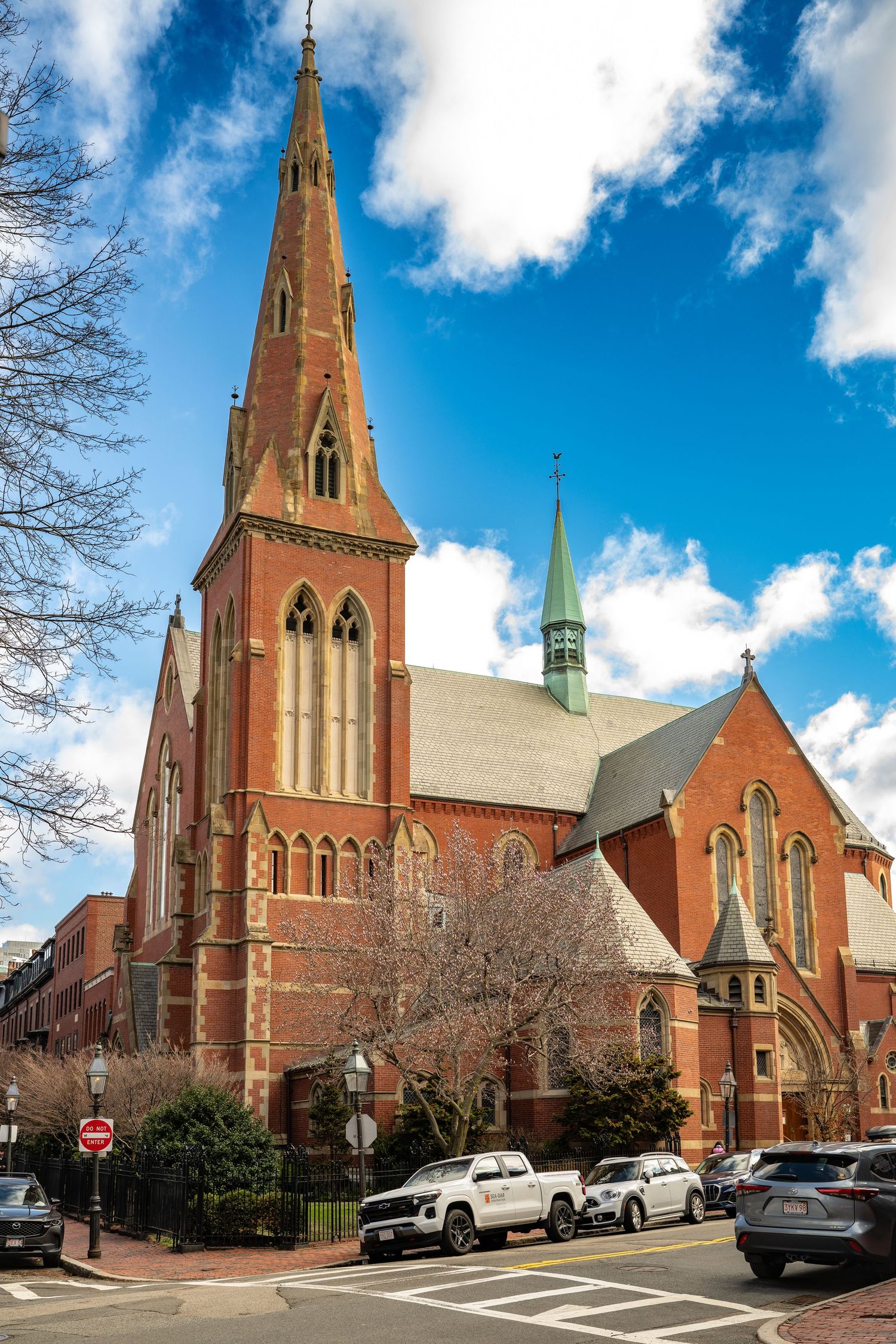 Gothic red brick church with tall spire under blue sky, cars parked on street in historic neighborhood.