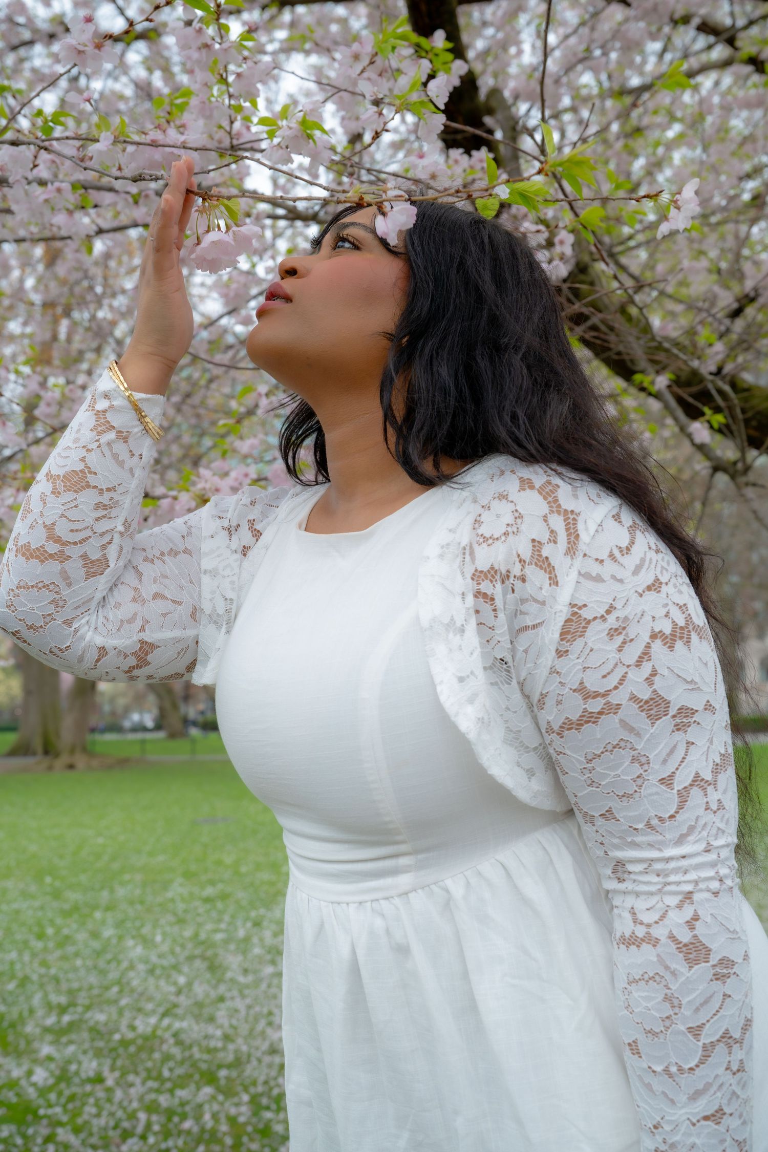 Woman in white lace dress reaches toward pink cherry blossom branches in a serene spring garden.