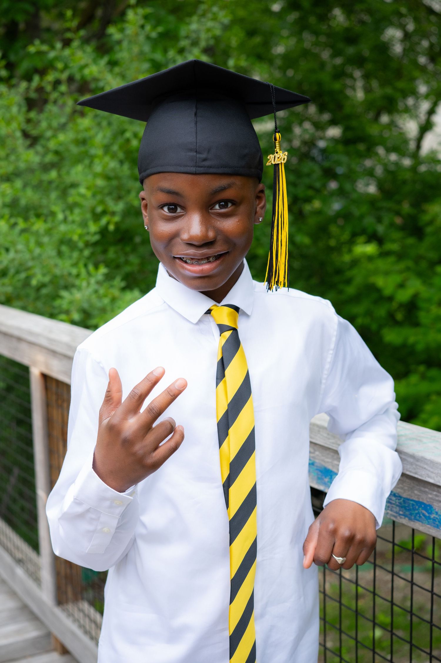 Smiling graduate in cap and yellow striped tie poses on outdoor bridge.
