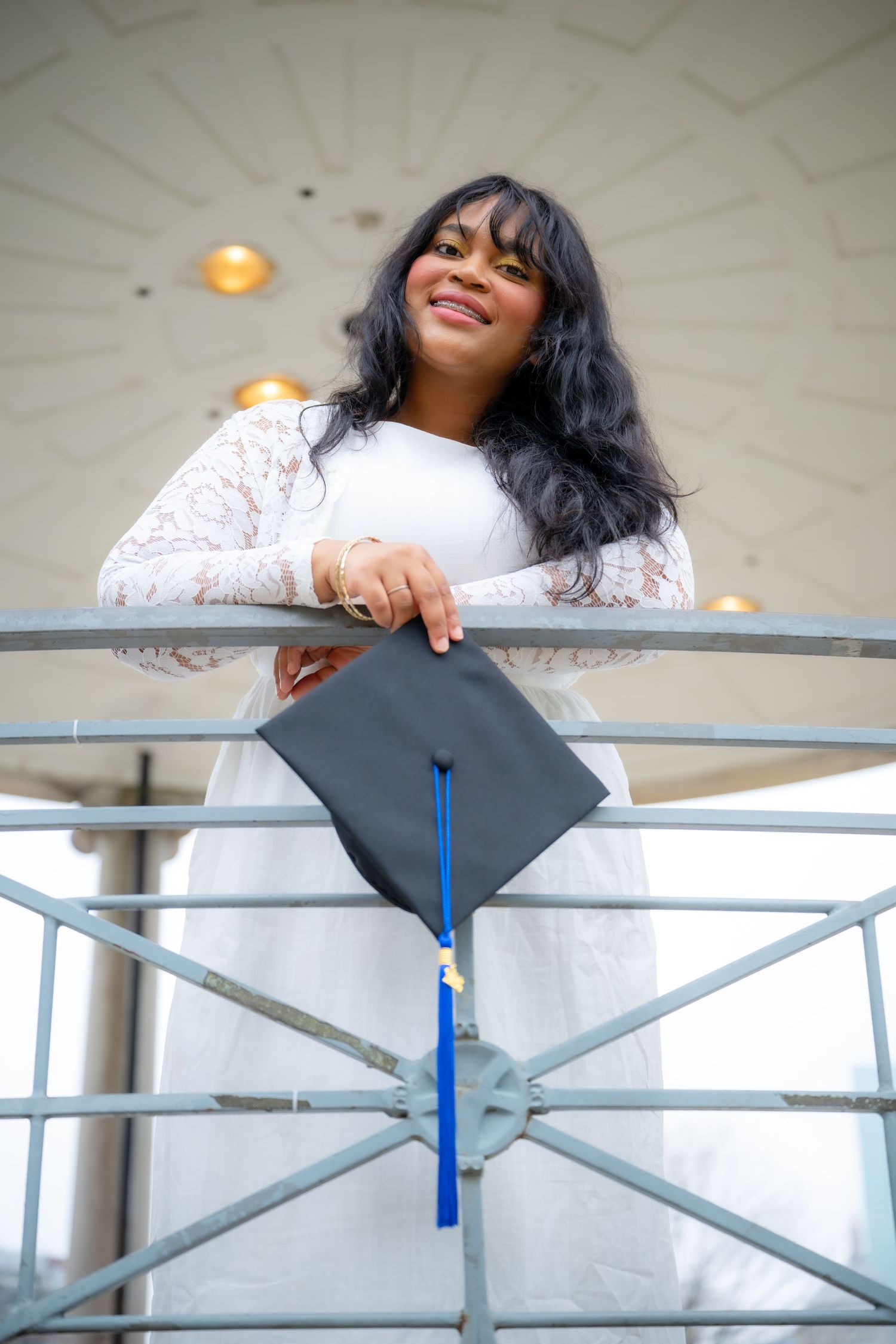Smiling graduate in white lace dress holding cap while leaning on a railing outdoors.