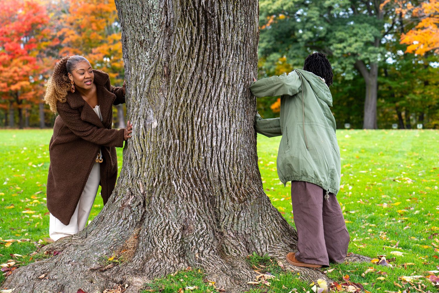 Two people in cozy jackets stand on opposite sides of a large tree trunk in a grassy park surrounded by colorful autumn foliage.