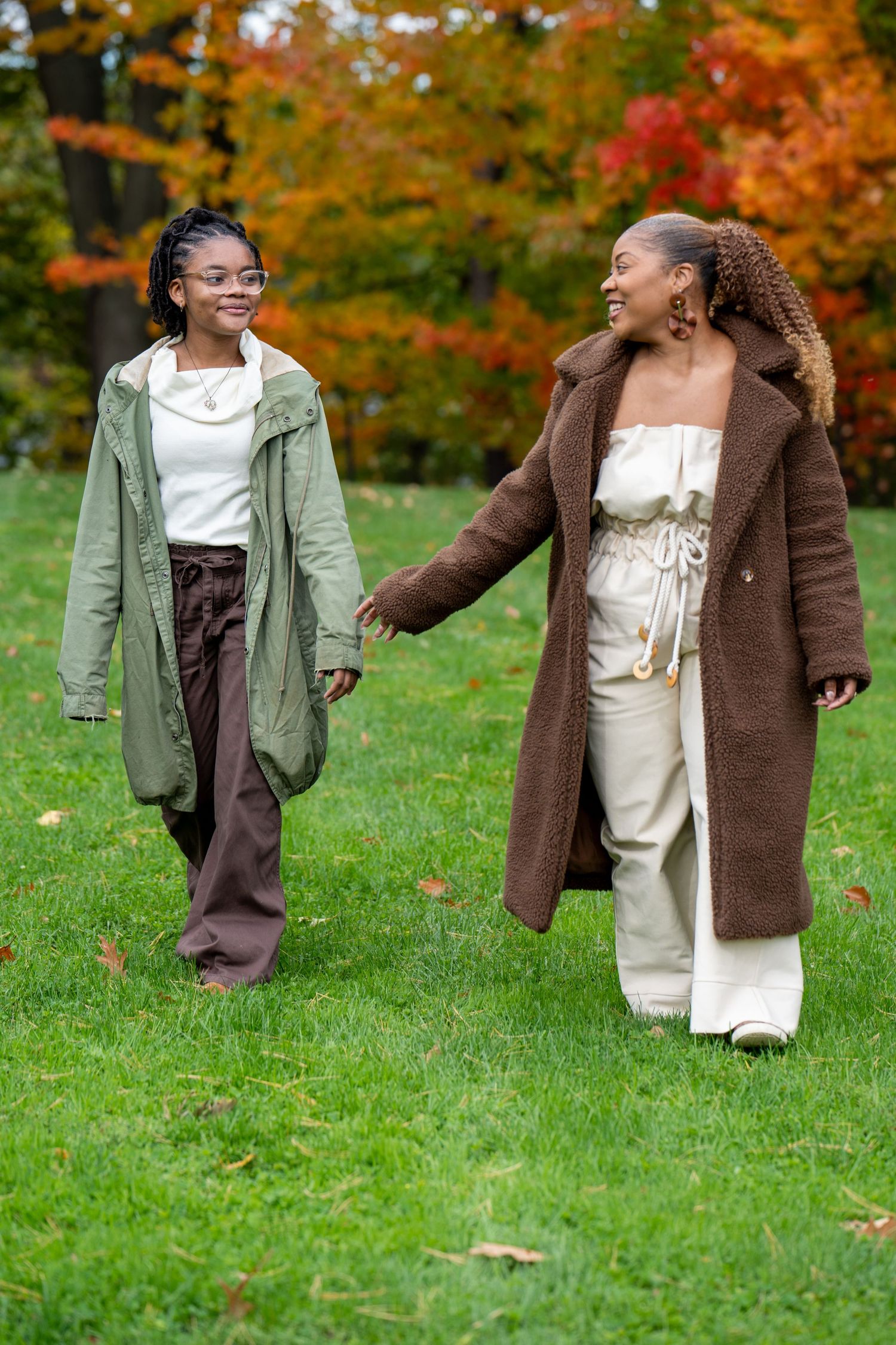 Two women walk hand in hand across a green lawn in a park, dressed in cozy fall layers with colorful autumn trees in the background.
