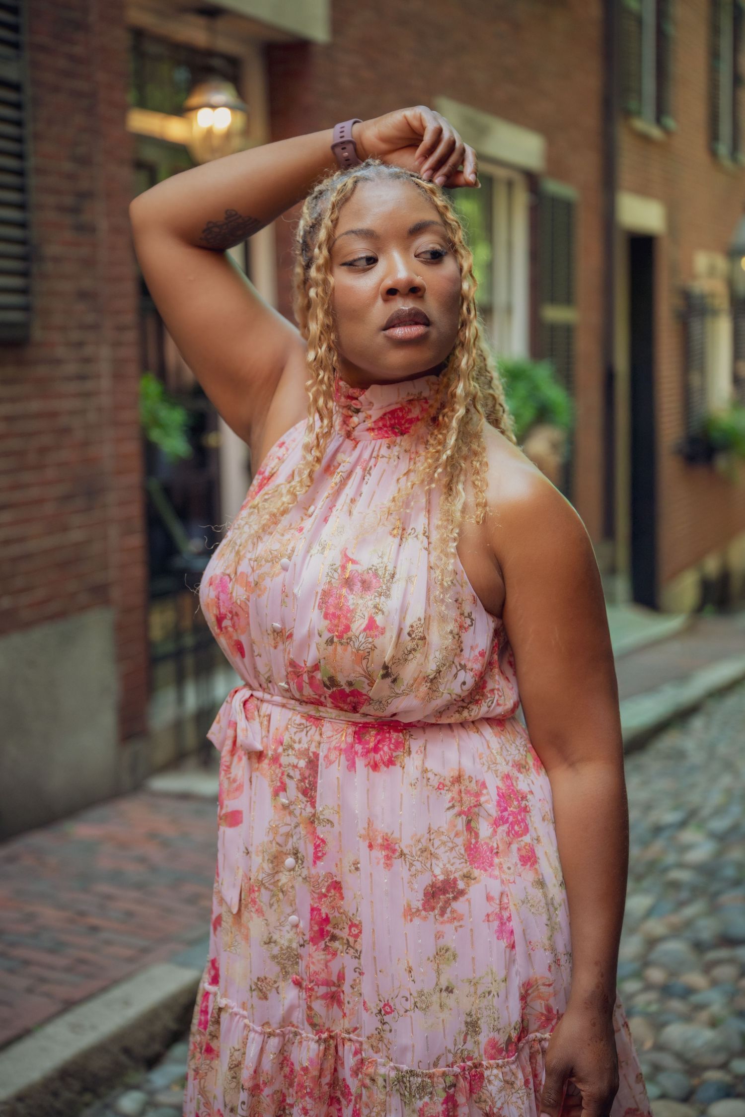 A woman in a floral pink halter dress poses confidently on a cobblestone street.