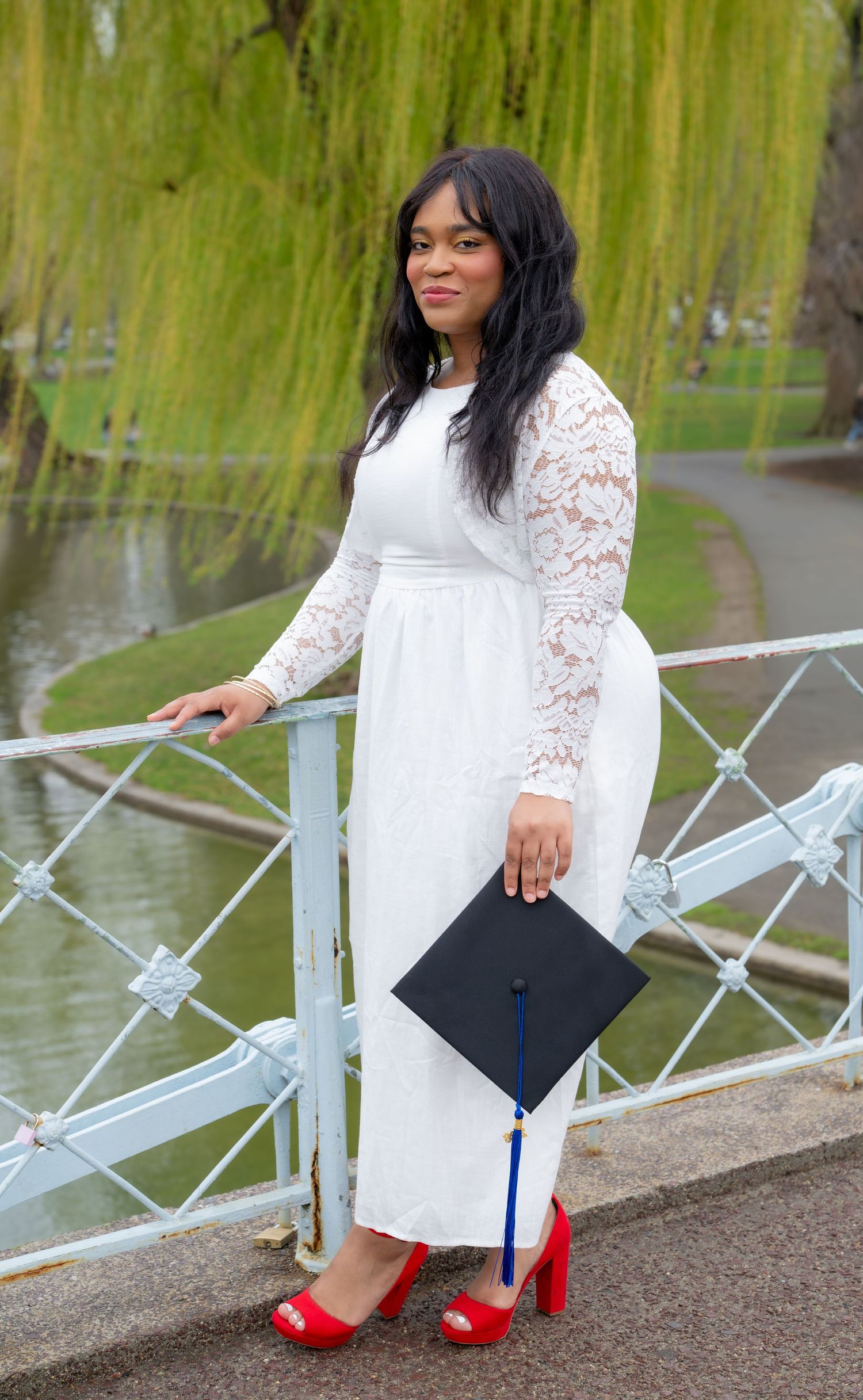 Graduate in white lace dress holding cap by a pond with willow trees in a park.