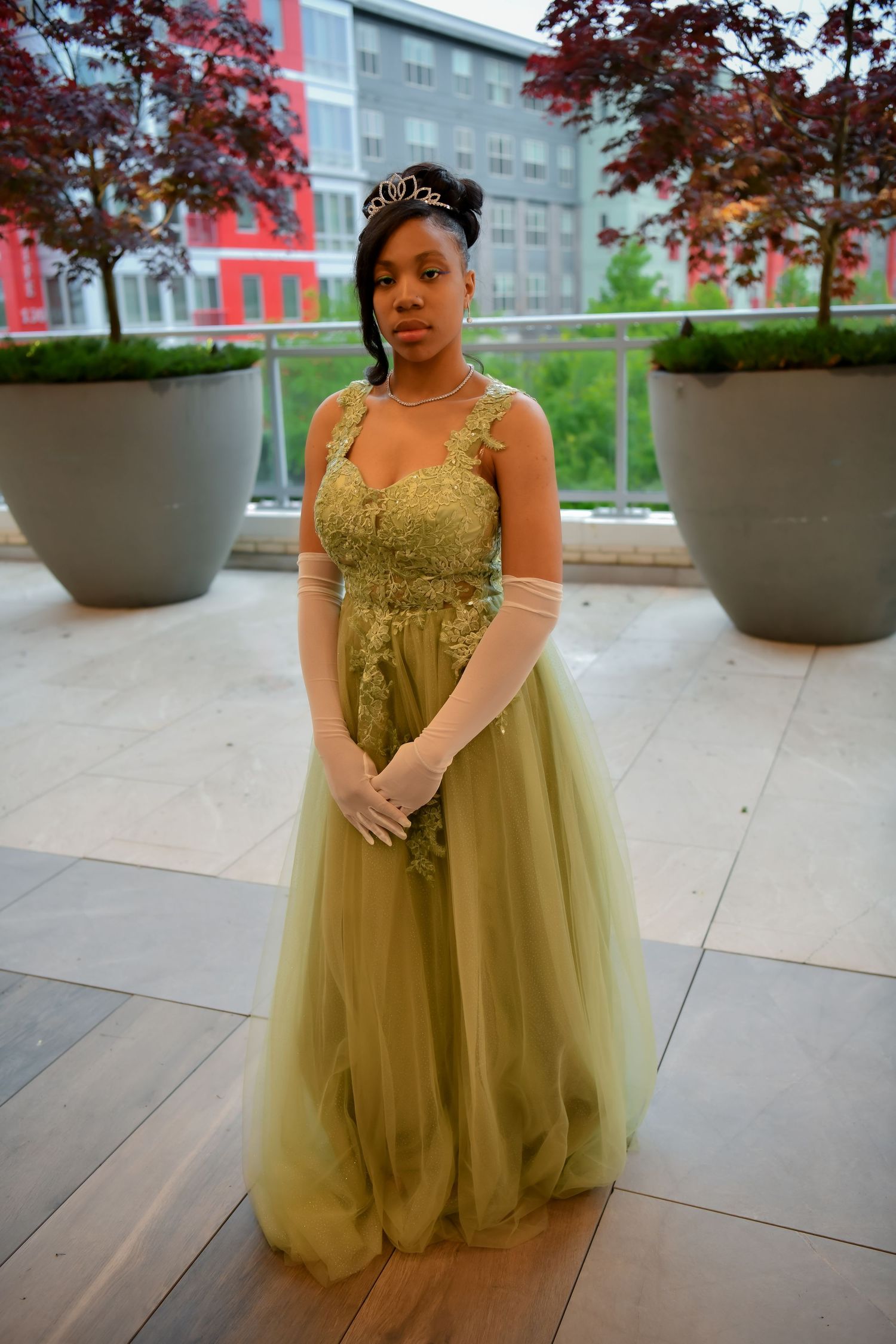 A young woman poses elegantly in a green lace ballgown with a tiara and white gloves.