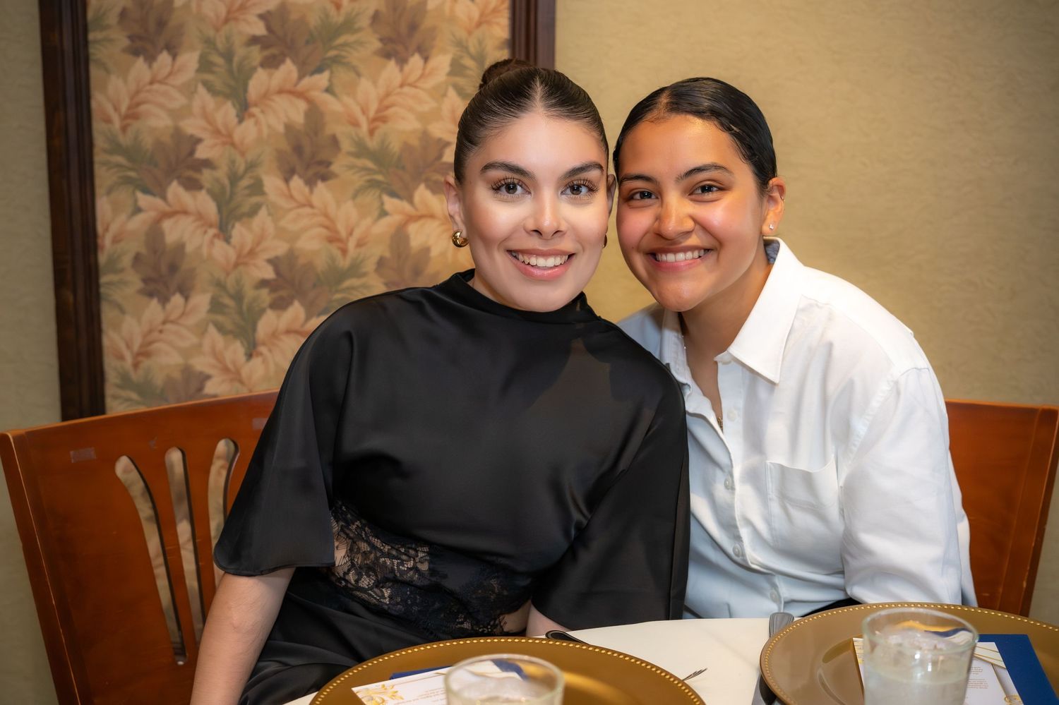 Two smiling young women pose together at a dining table in a warmly decorated restaurant.