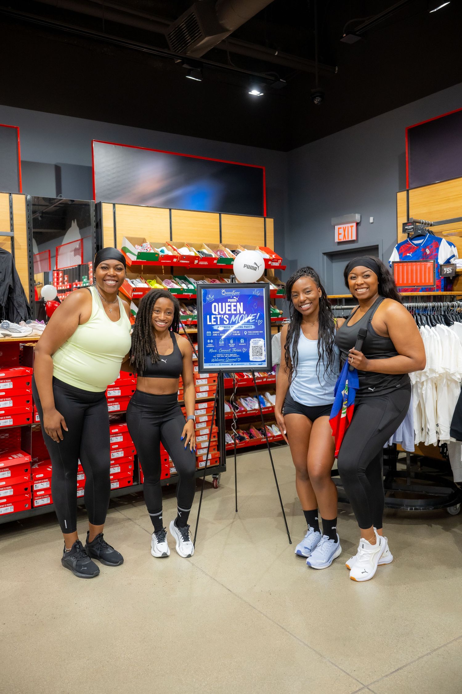 Four women in athletic wear pose together inside a Nike store near a Queen promotional display.