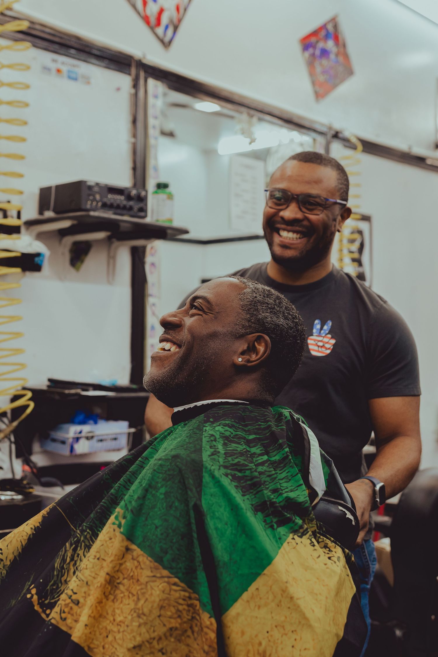 A barber and client share a laugh in a barbershop, client draped in a Jamaican flag cape.