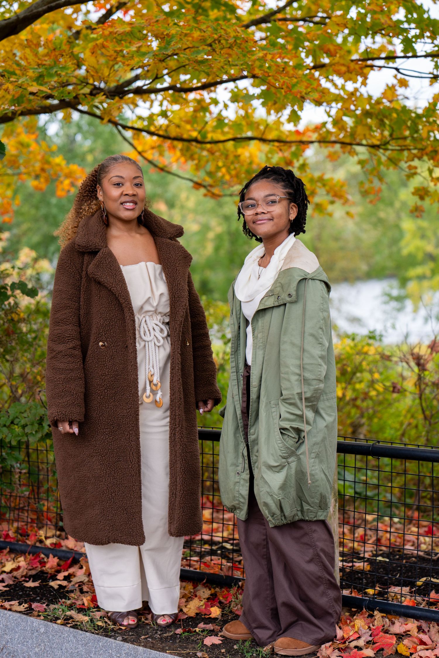 Two women in cozy fall coats, one brown and one olive green, stand by a railing beneath trees with colorful autumn foliage.