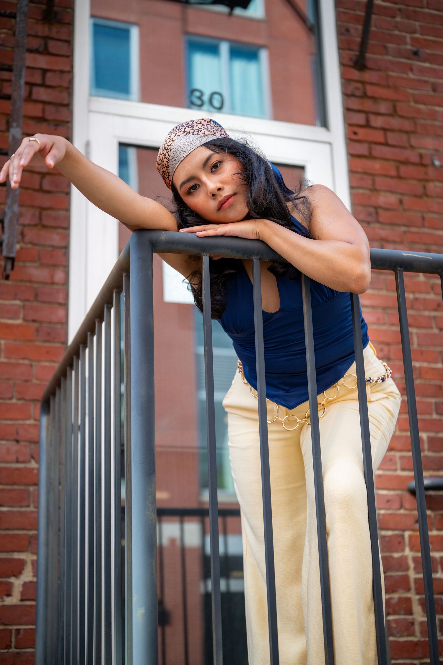 Young woman in blue top and cream pants leans on metal railing by brick building.