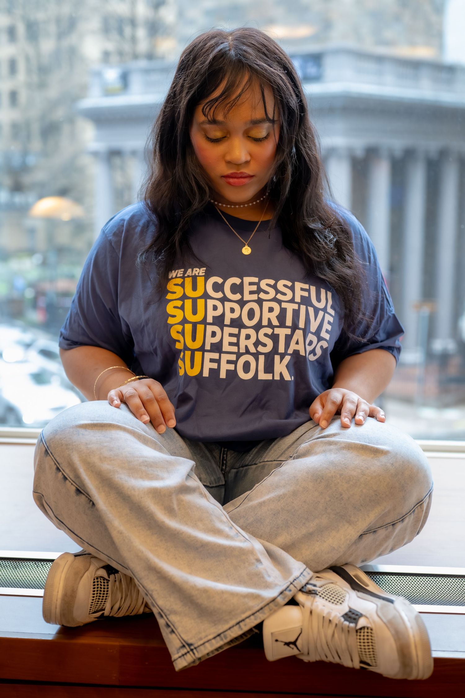 Young woman in Suffolk University t-shirt sits cross-legged on window ledge.