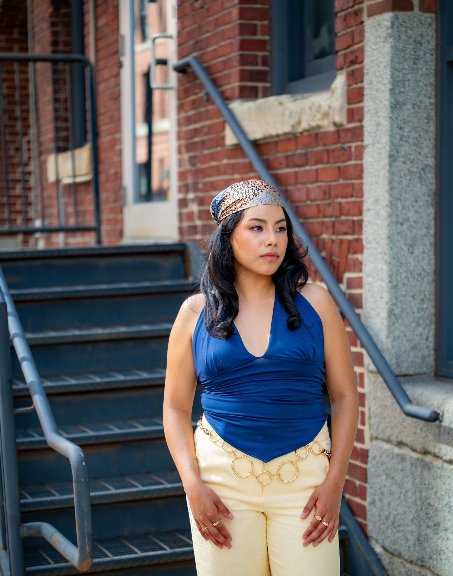 Woman in blue halter top and yellow pants with head wrap poses on urban brick building steps.