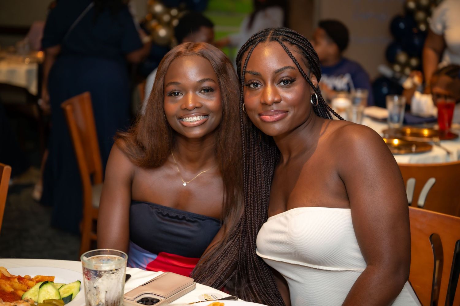 Two young women smile together at dinner table during festive indoor graduation celebration event.