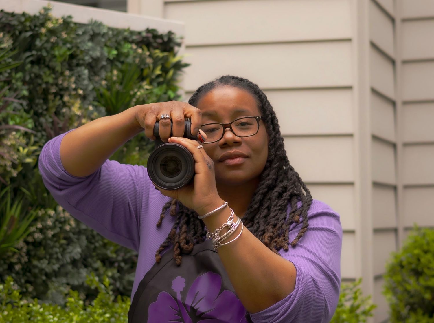A young Black woman with braids and glasses holds a DSLR camera up to photograph outdoors near green foliage.