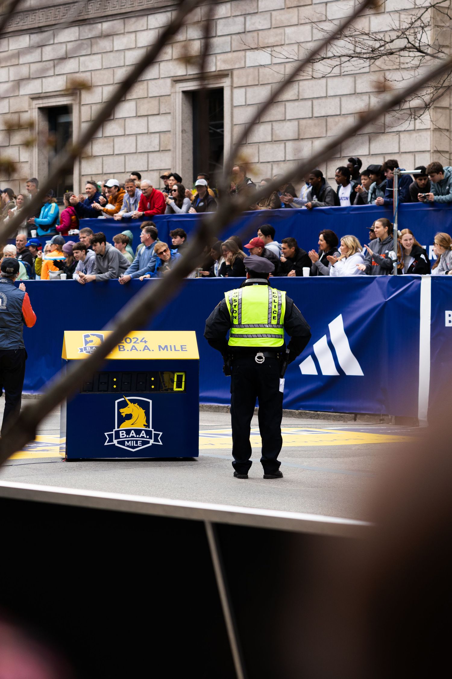 Security officer in hi-vis vest stands at BAA mile marker during Boston Marathon race event with spectators.
