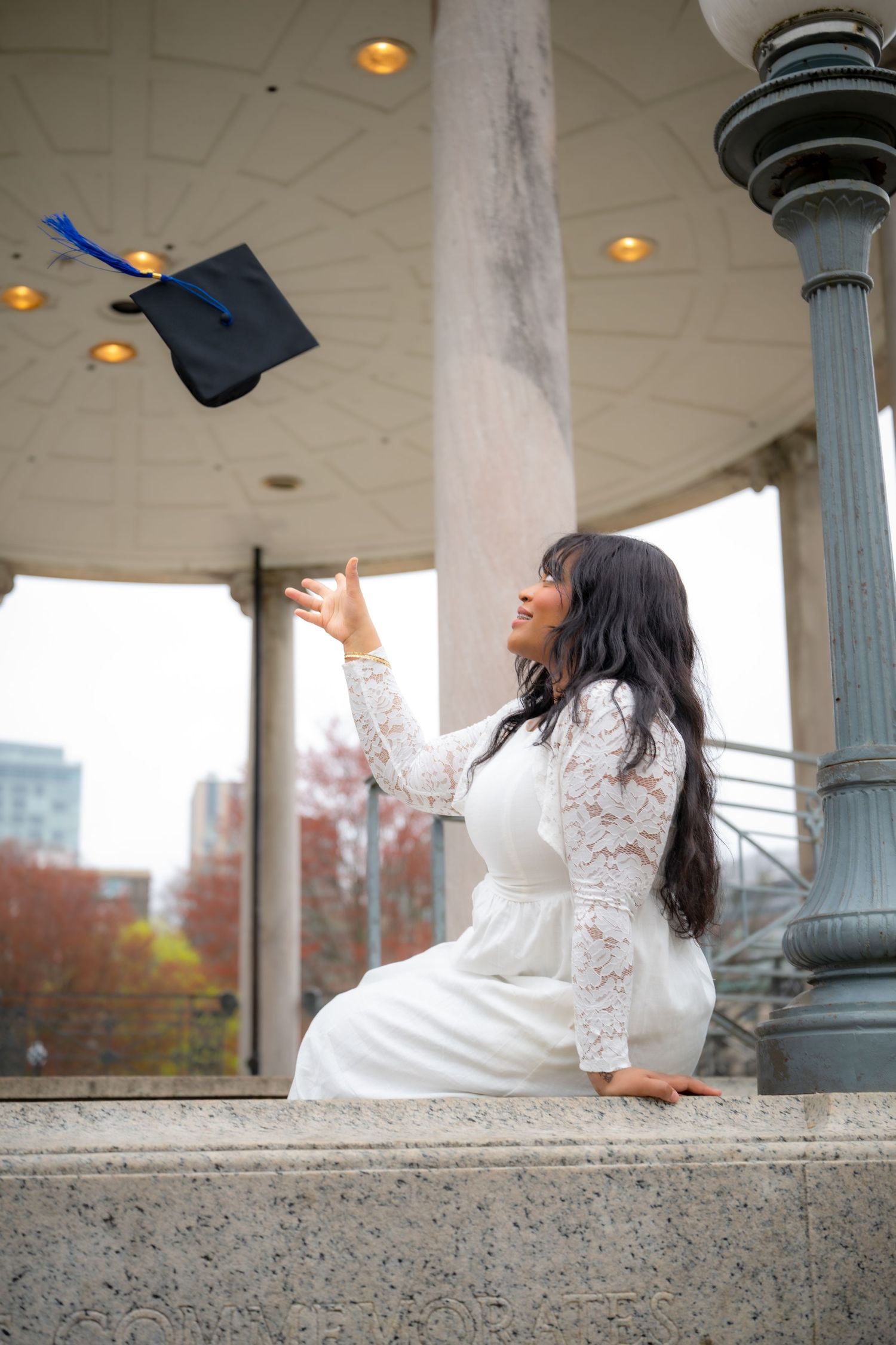Graduate in white lace dress tossing cap in the air at an outdoor pavilion.