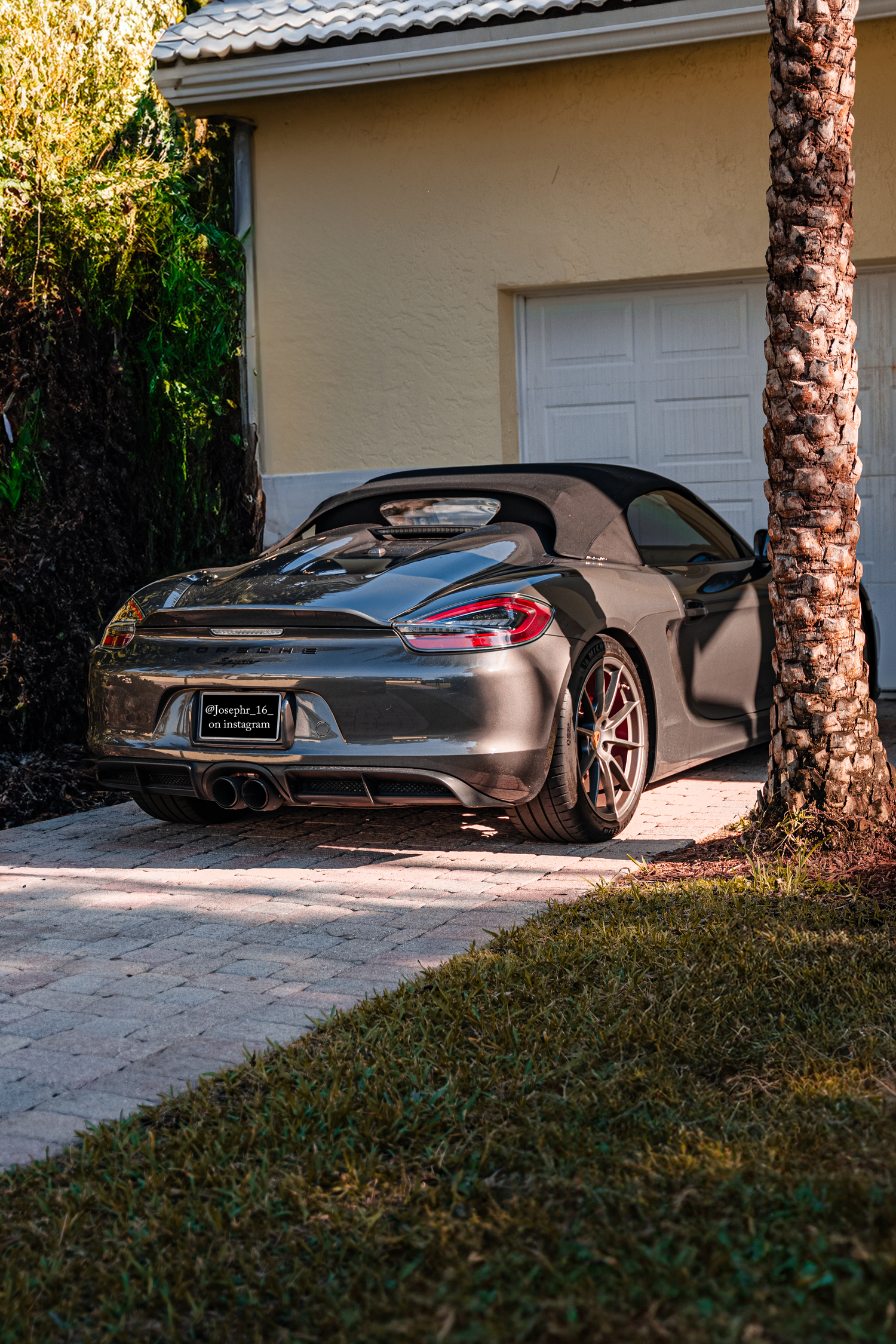 Silver Porsche Boxster convertible parked in a residential driveway beside a palm tree and white garage.