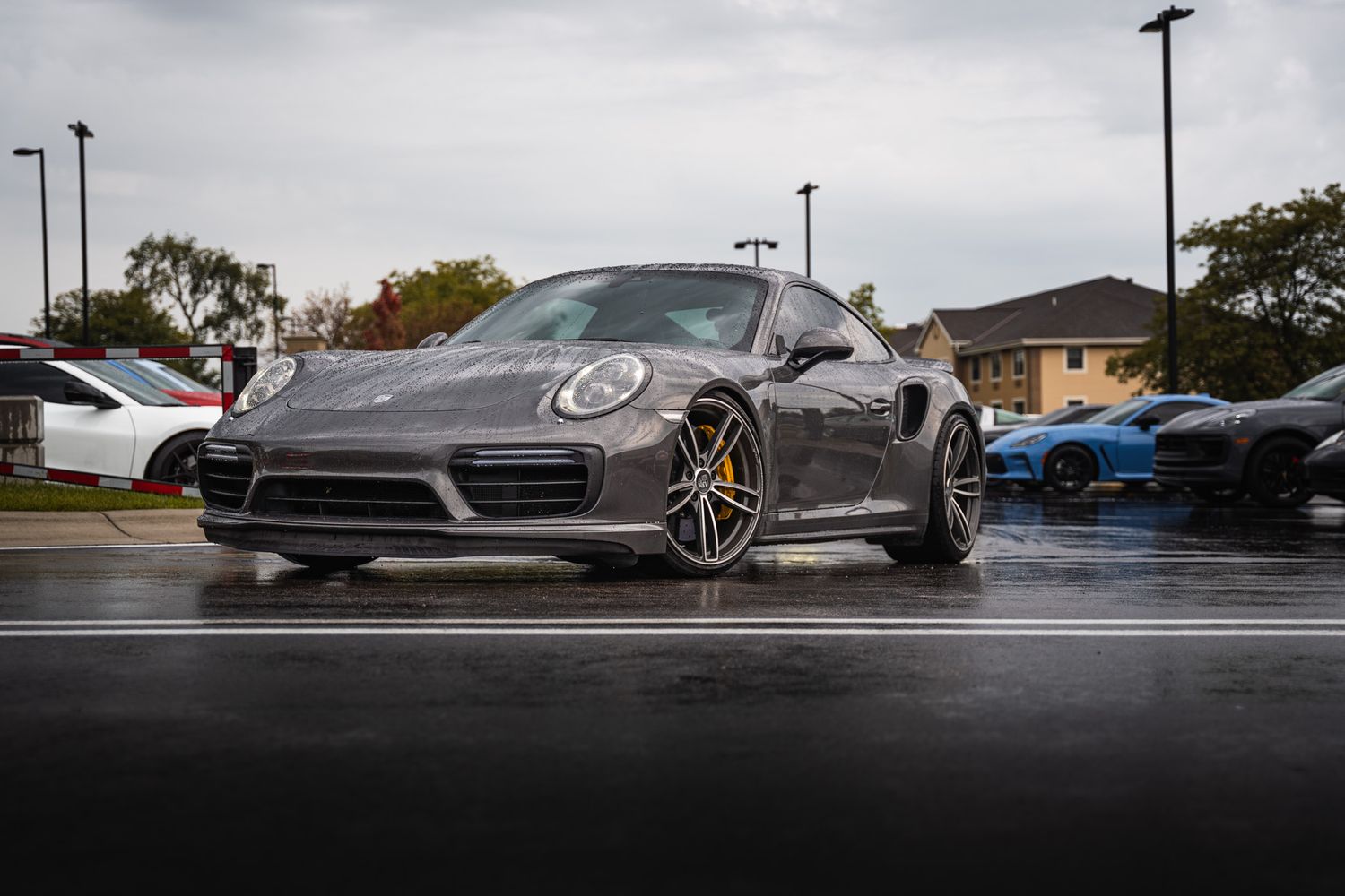 Gray Porsche 911 Turbo S coupe parked on wet pavement at a car meet in a parking lot.