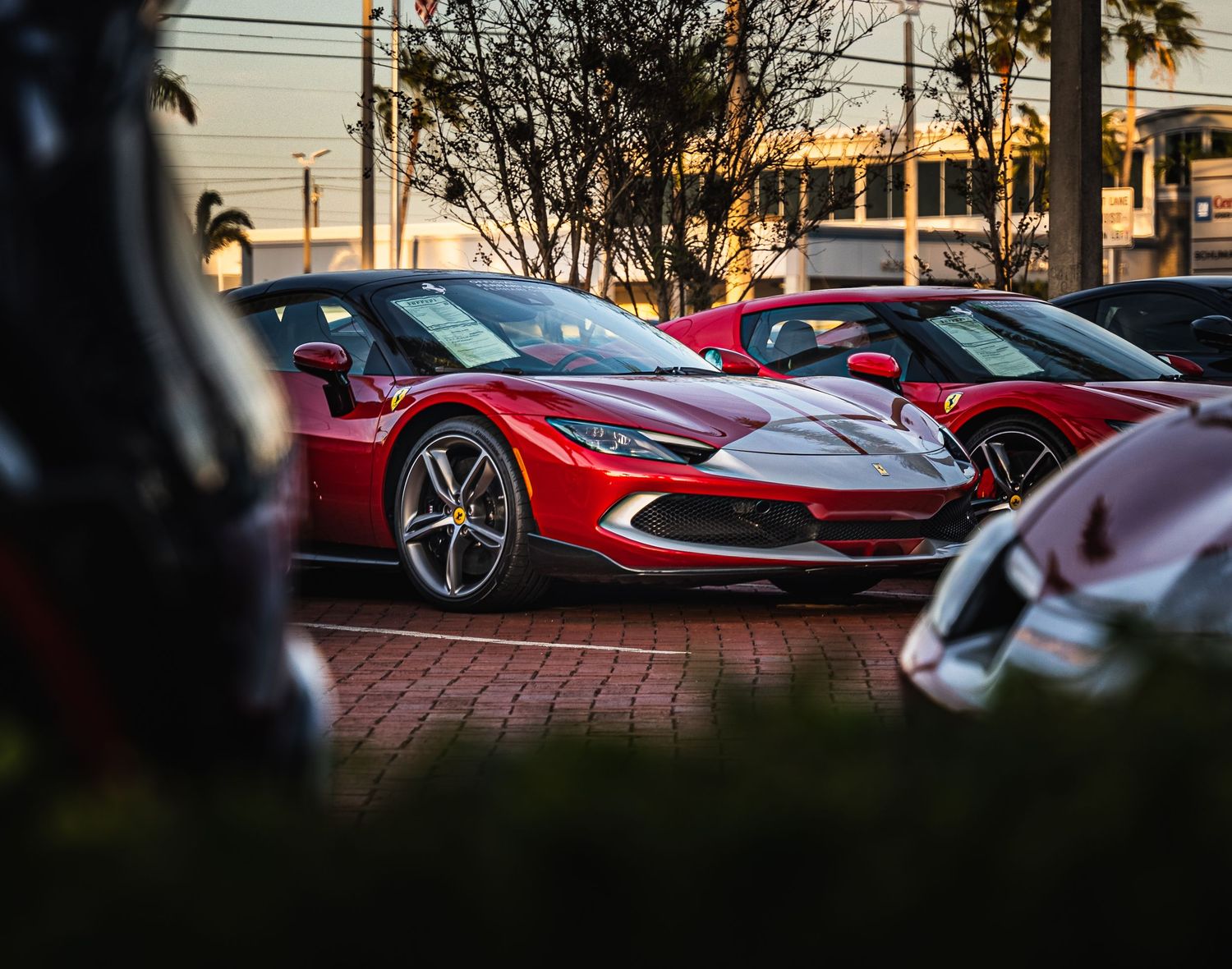 Red Ferrari 296 GTB supercar parked at a sunset car meet surrounded by exotic vehicles.