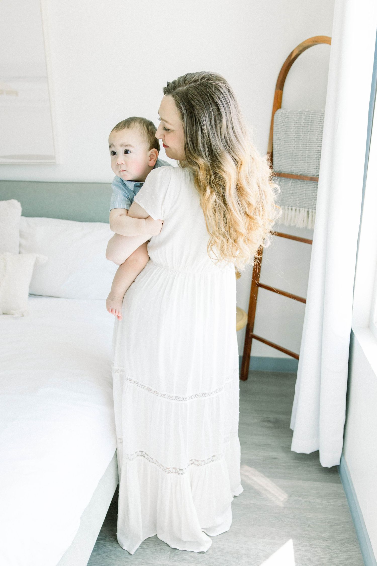 Mother in a white dress holding her baby by a window in a soft, natural-light bedroom during a San Diego motherhood session