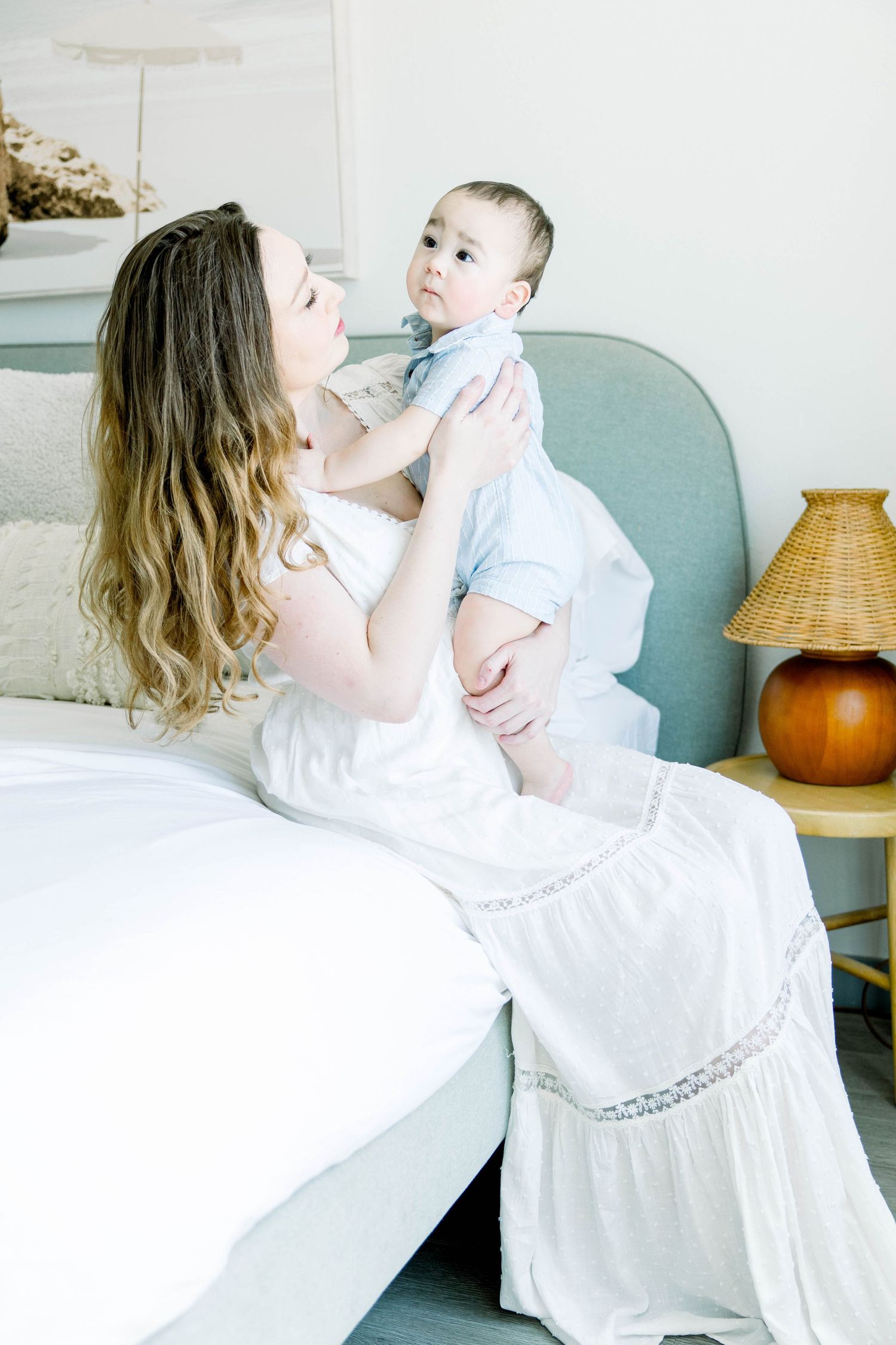 Mother in a flowing white dress lifting her baby while sitting on the edge of a bed during a San Diego in-home session