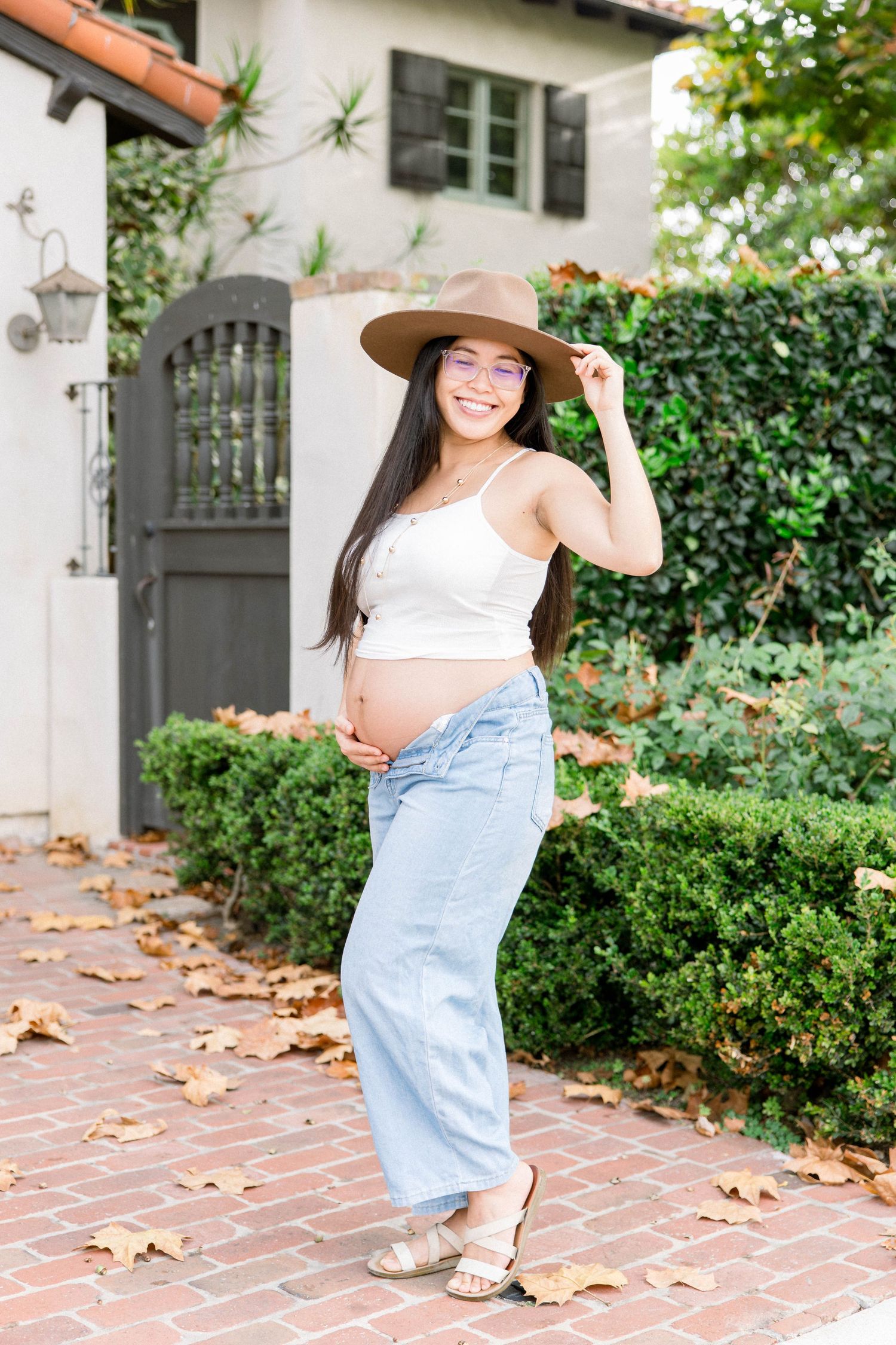 Pregnant woman in a tan hat smiling by a garden gate on a brick path during a San Diego maternity session