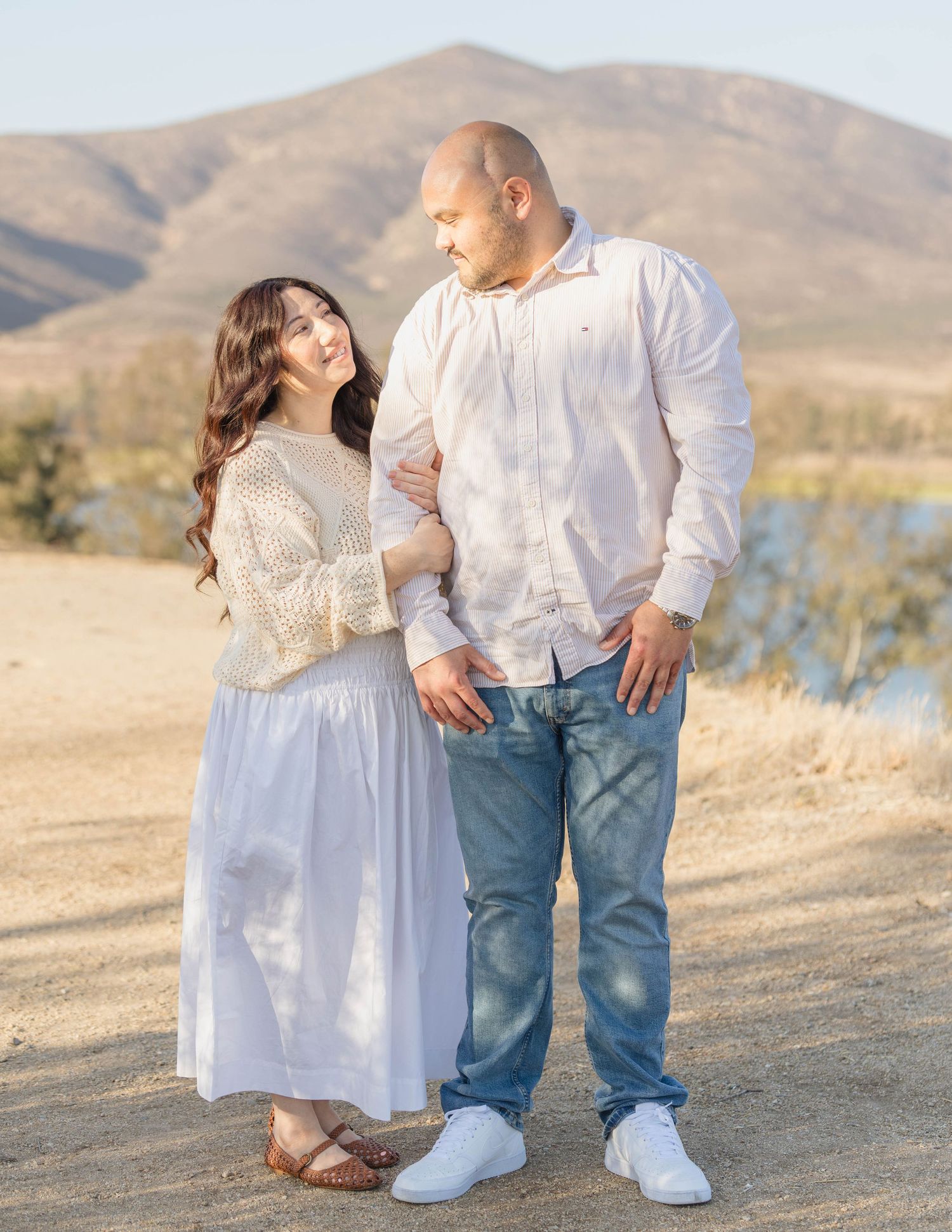 Couple in cream and white outfits standing close together with a mountain and lake behind them during a San Diego couples session