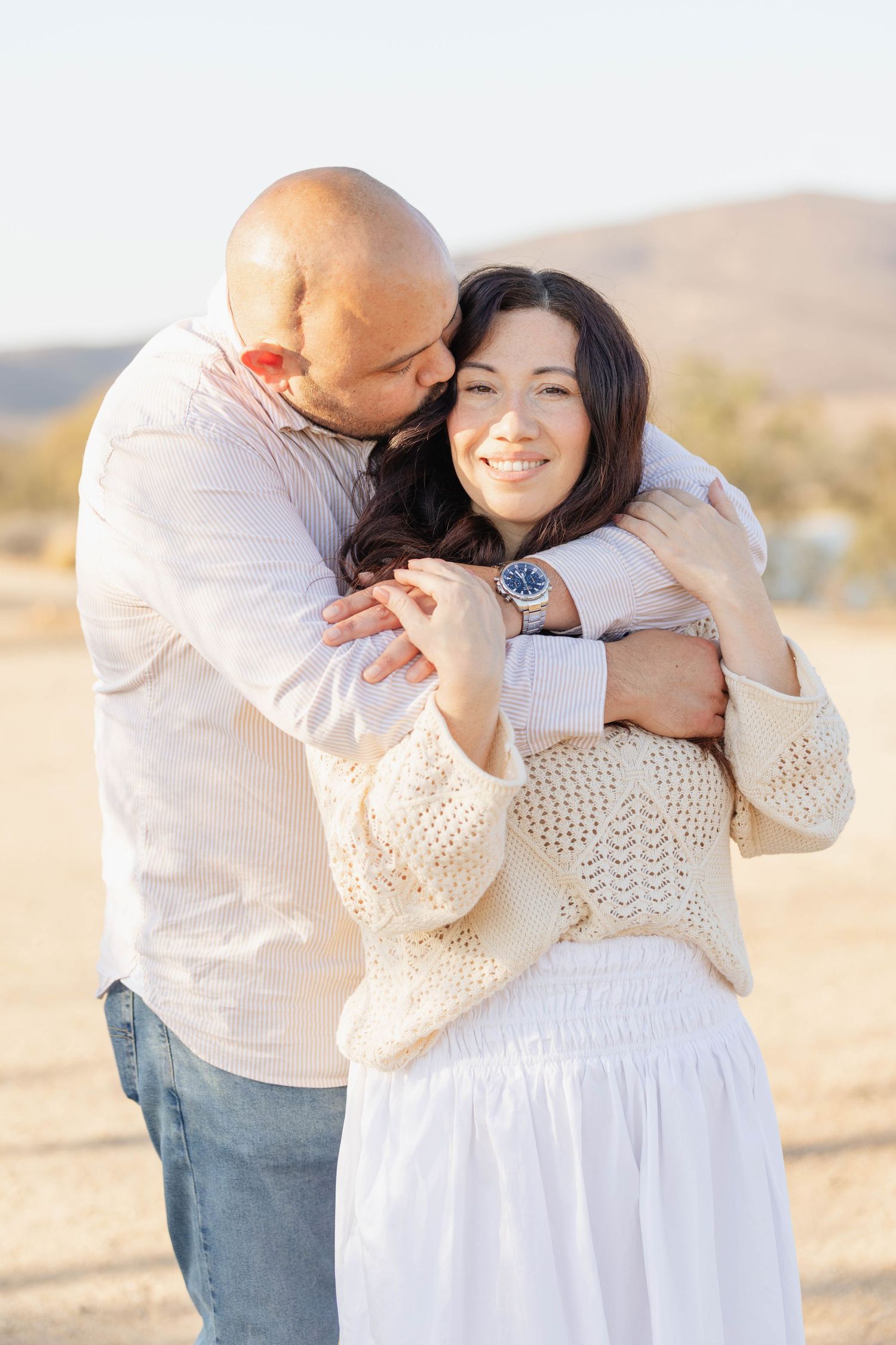 Smiling woman wrapped in her husband’s arms as he kisses her head during a golden hour couples photoshoot in San Diego