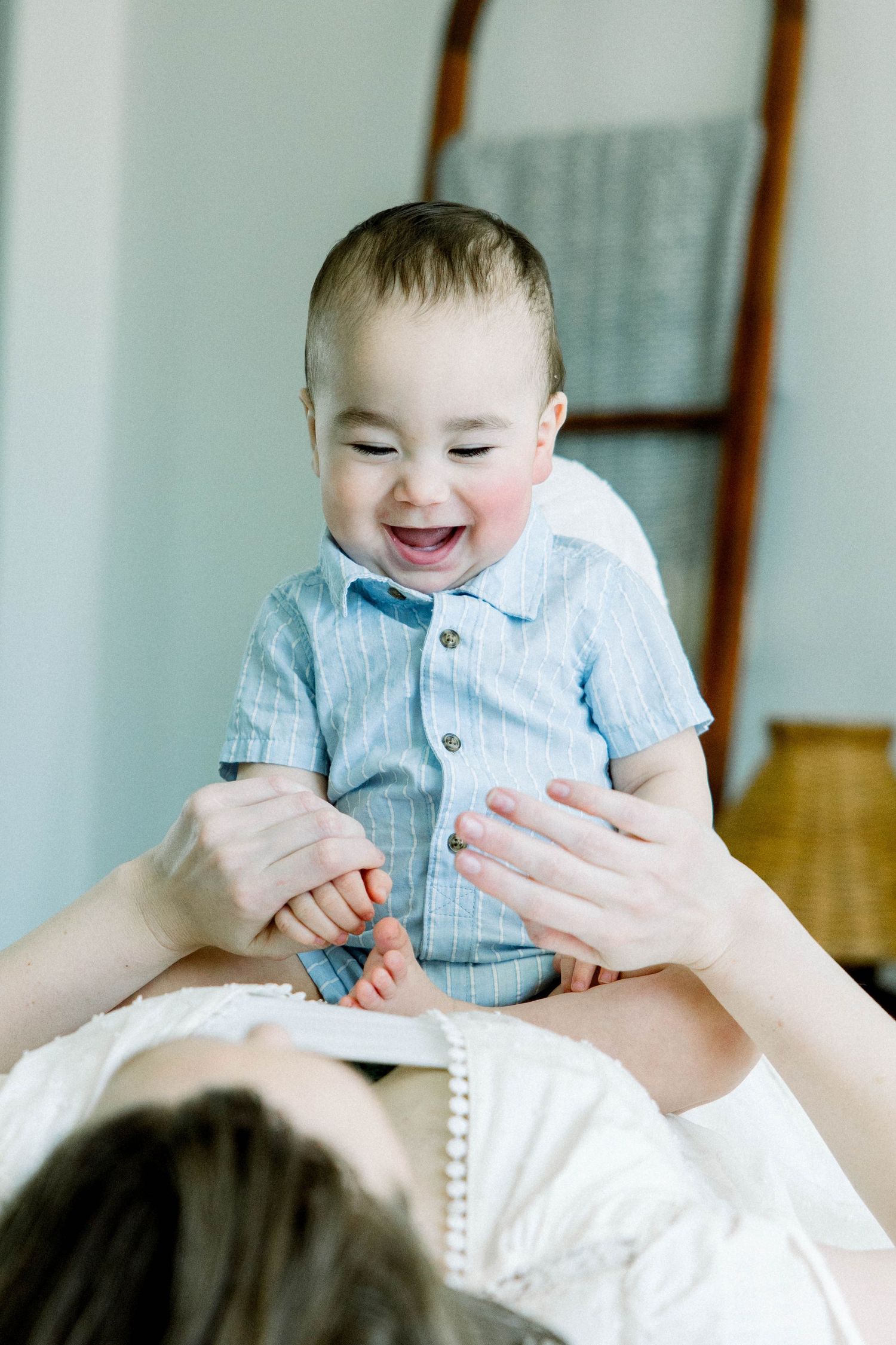 Happy baby in a blue button-up shirt sitting on his mother’s lap in a light, airy bedroom