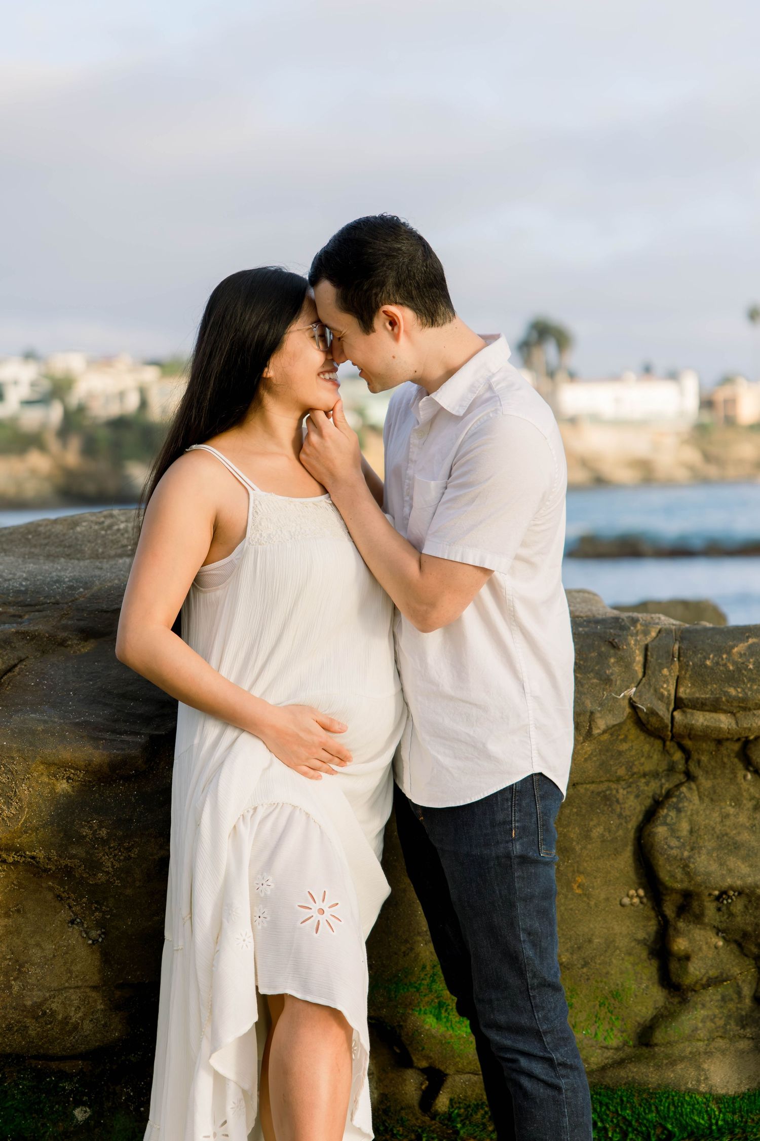 Expecting couple sharing a nose-to-nose moment on coastal rocks with the ocean behind them during a San Diego beach maternity session
