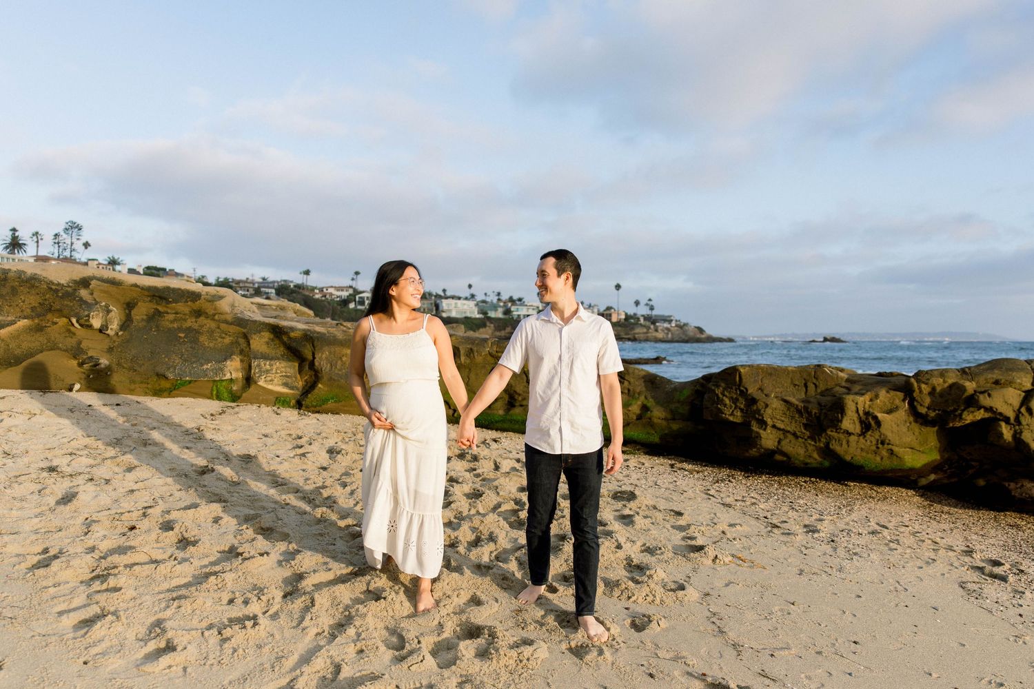Pregnant woman and her husband walking hand in hand on the beach at sunset during a San Diego maternity photoshoot