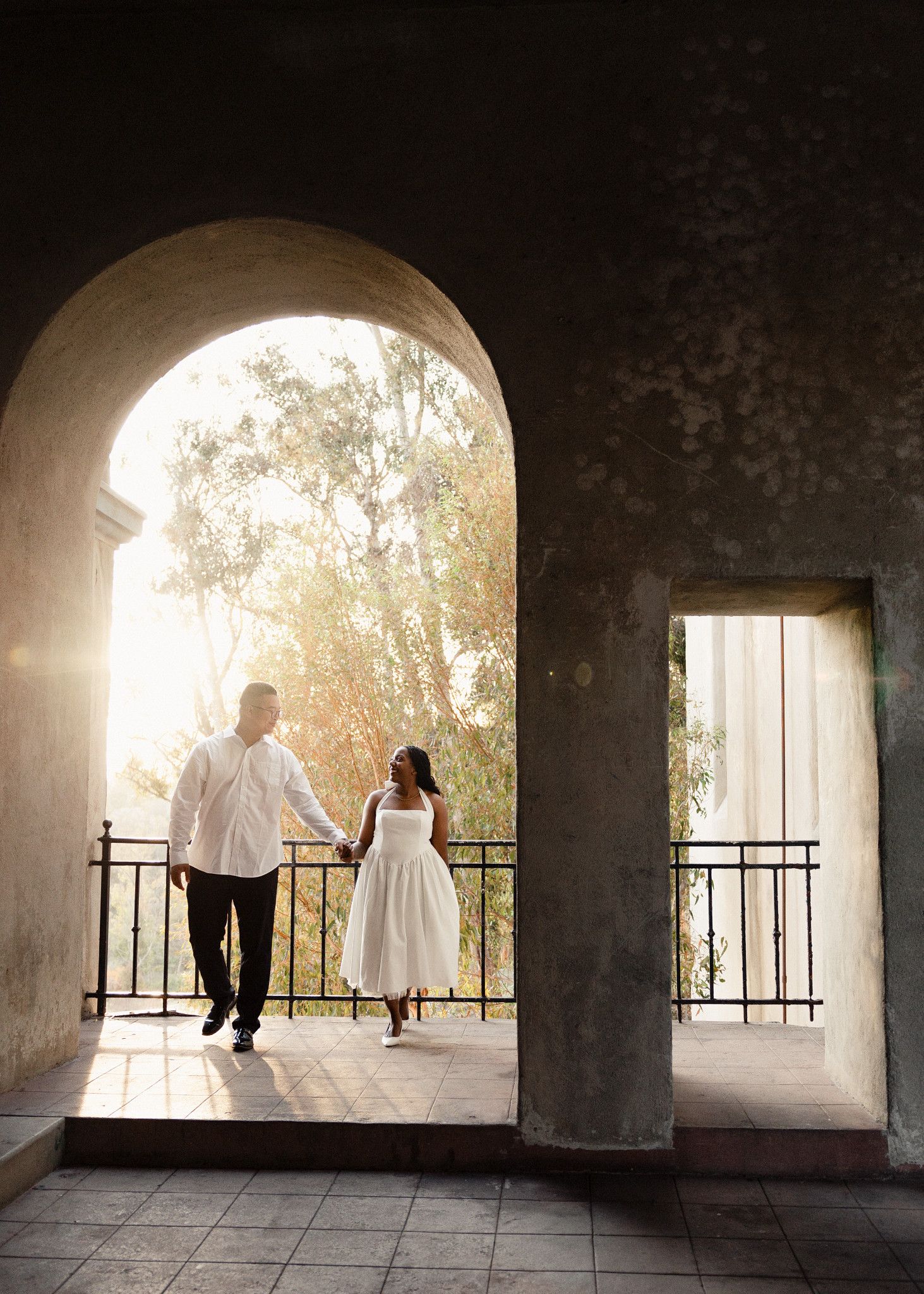 Couple holding hands walking out from under a stone arch at sunset in San Diego