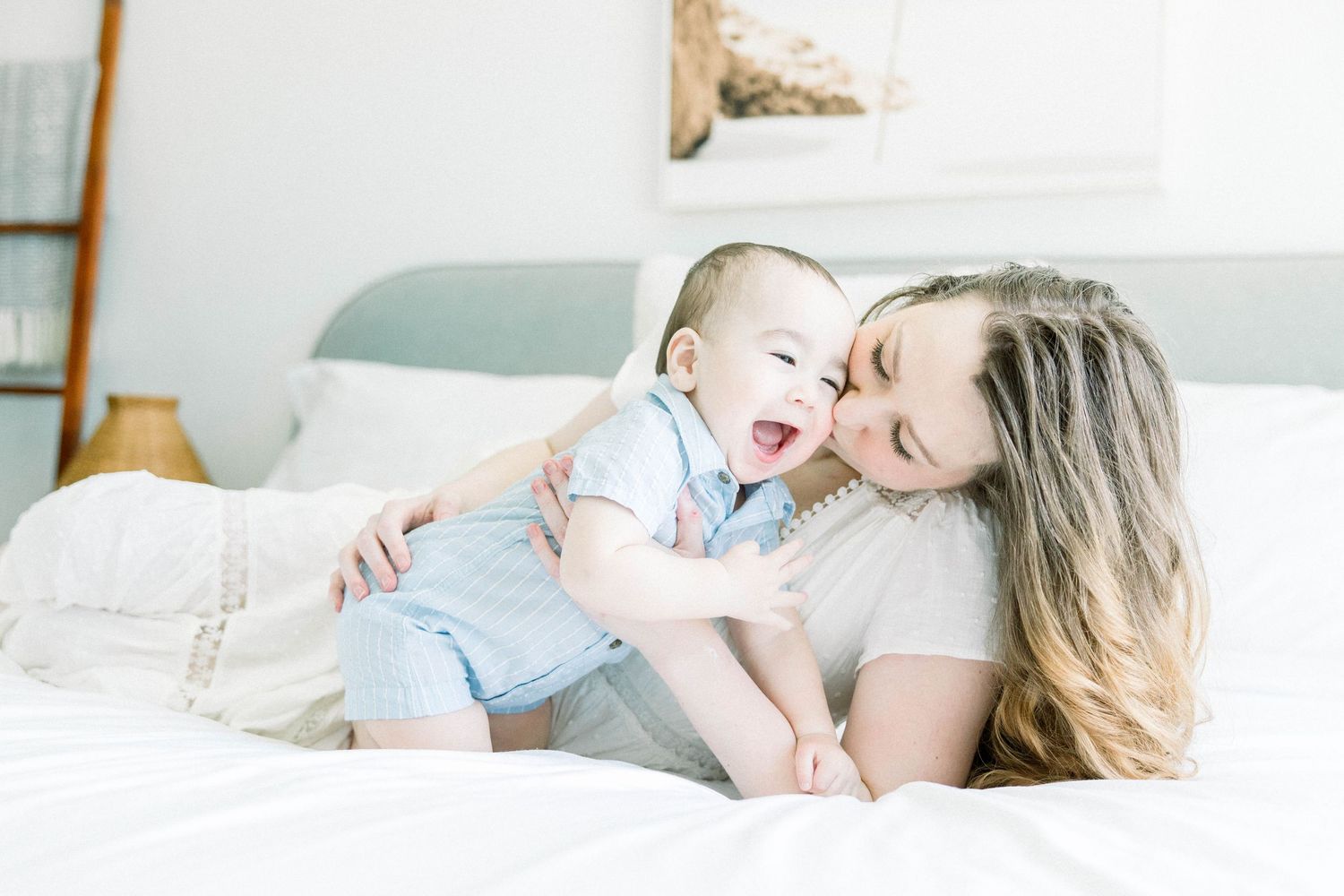 Mother nuzzling her laughing baby on a white bed during a bright, airy in-home motherhood session in San Diego