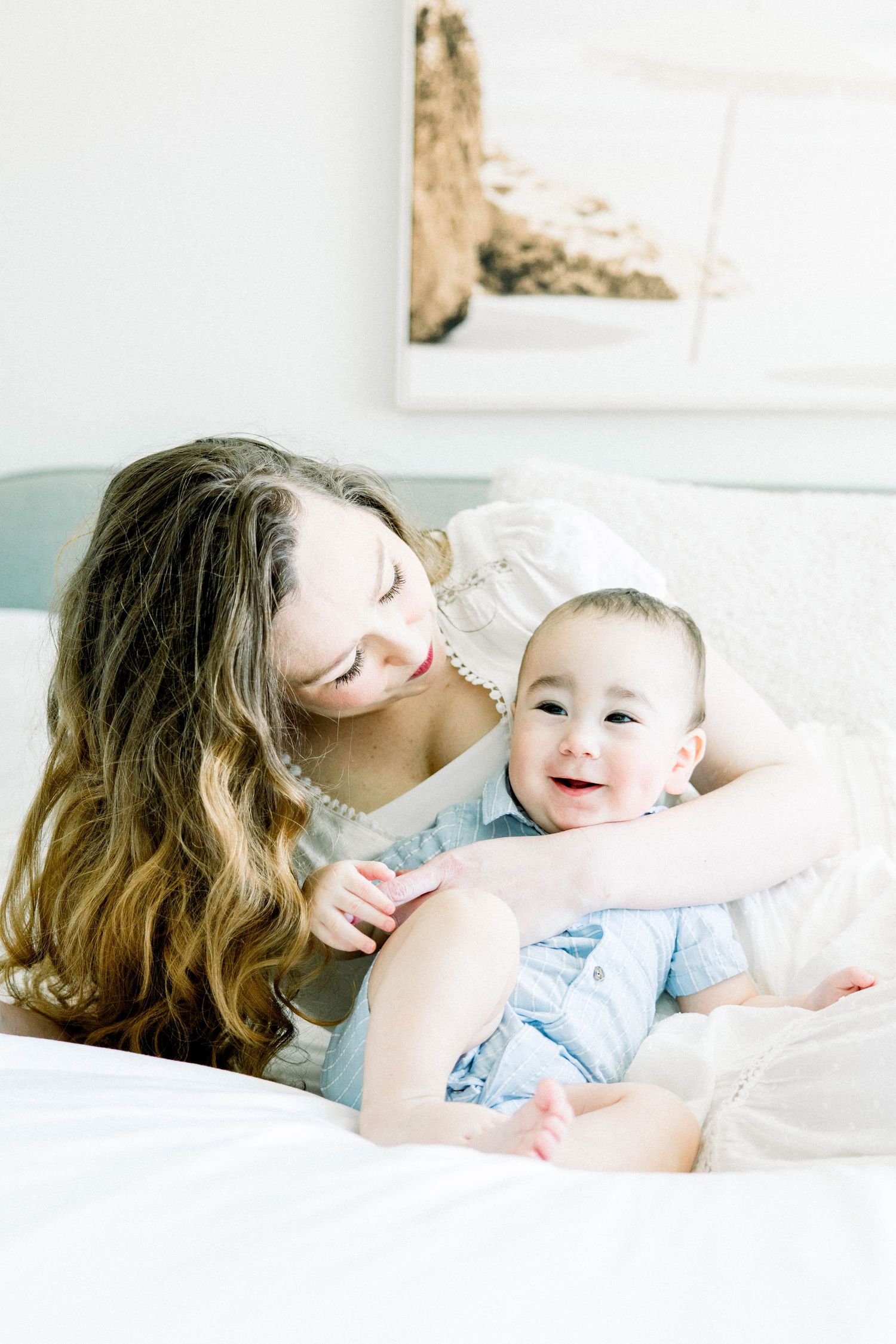 Smiling mother hugging her baby boy on a bed with soft natural light during a San Diego family lifestyle session